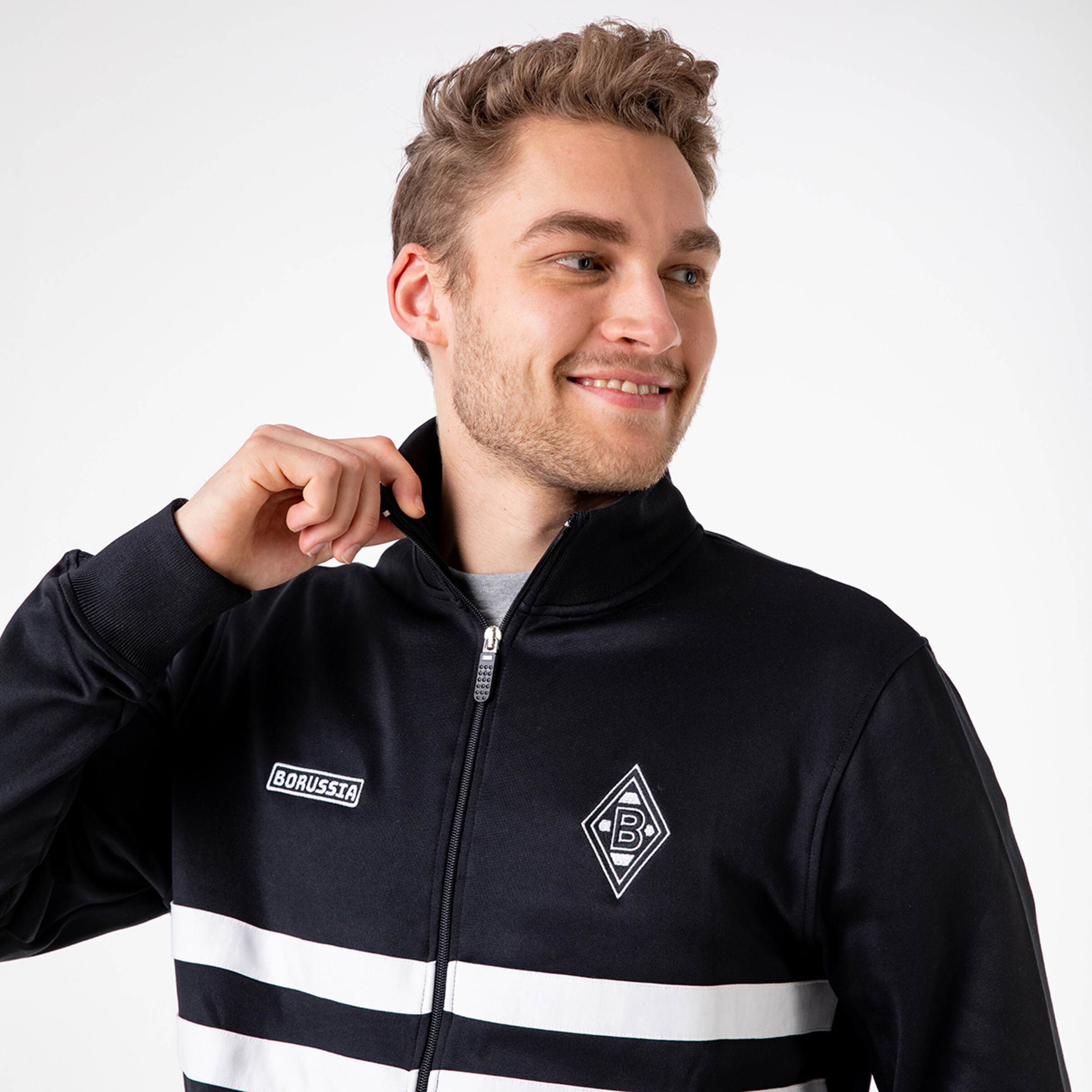 Mann in schwarzer Sportjacke mit weißen Streifen lächelt und greift an den Reißverschluss.