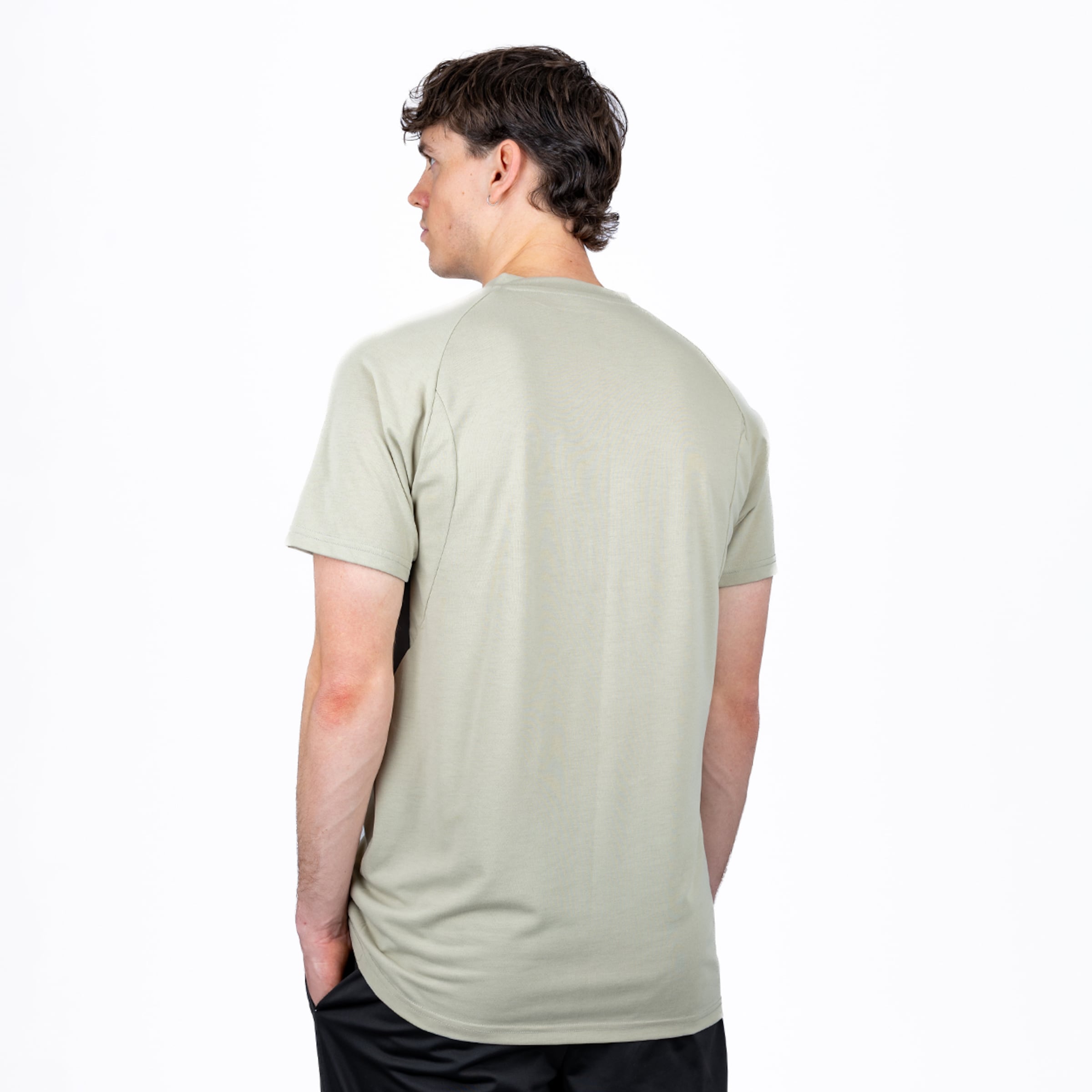 Ein Mann von hinten, blickt nach links über die Schulter, trägt ein hellgrünes T-Shirt mit Raglanärmeln und schwarze Shorts, Hände in den Taschen. Weißer Hintergrund.