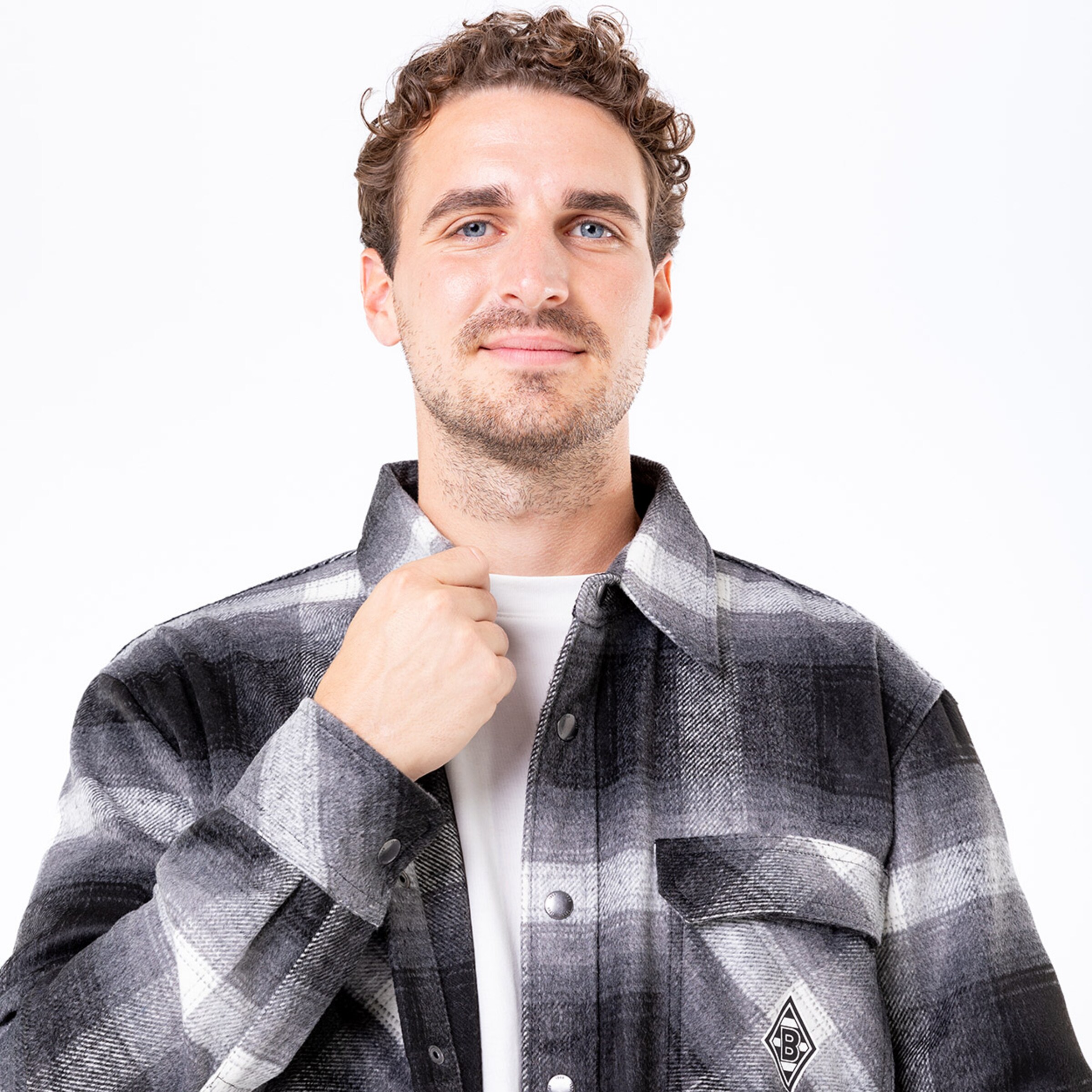 Mann mit lockigen Haaren in einem karierten Hemd und T-Shirt lächelt und fasst an den Kragen.