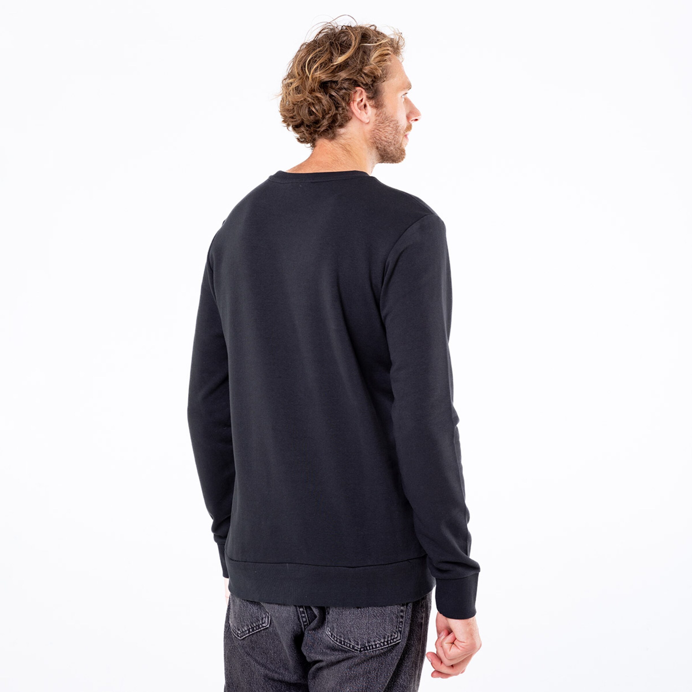 Ein Mann trägt einen schwarzen Pullover und steht mit dem Rücken zur Kamera. Er hat lockige Haare und trägt graue Jeans.