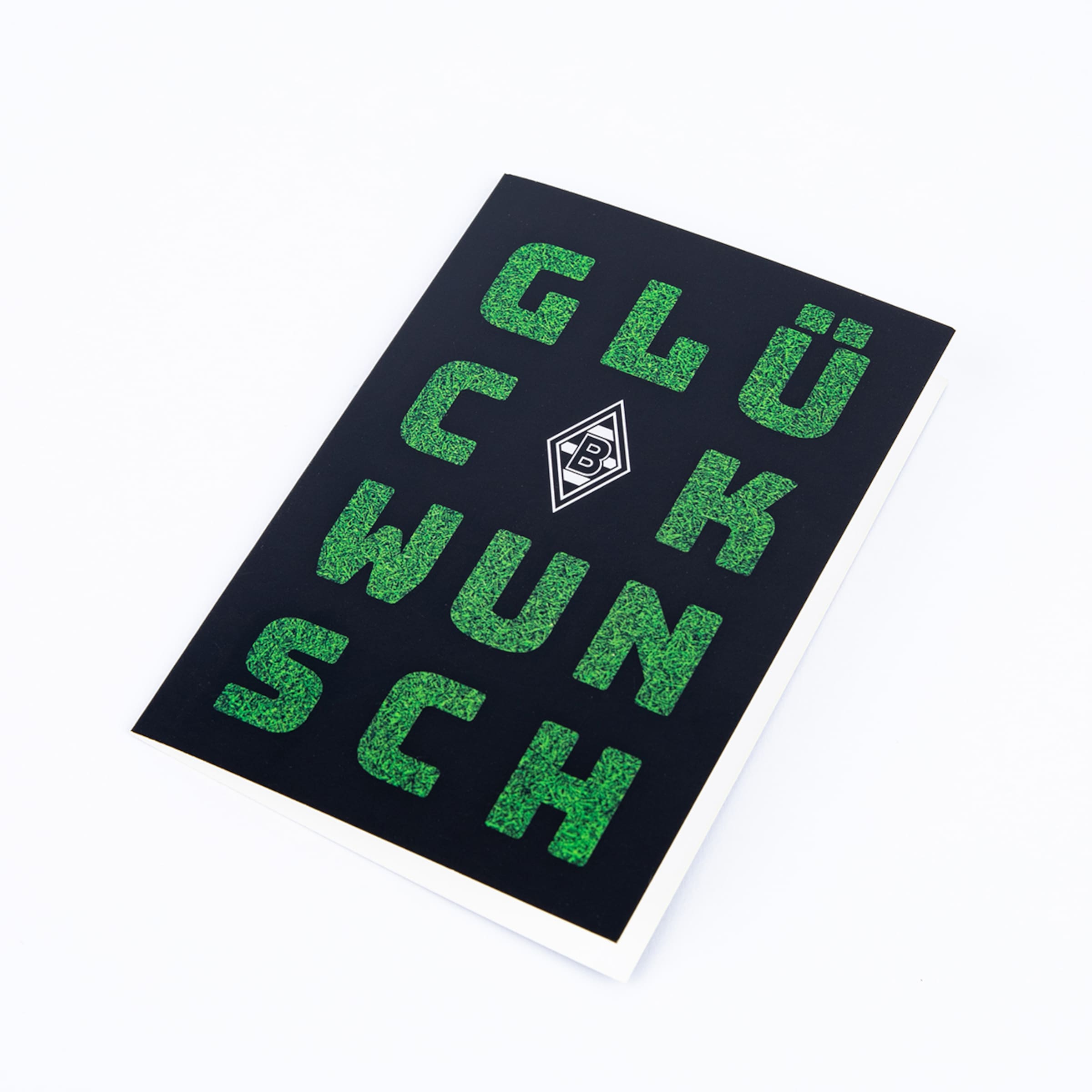 A black greeting card with 'GLÜCK' and 'WUNSCH' stacked vertically in green, grass-textured letters, separated by the Borussia Mönchengladbach logo. The card lies diagonally on a white background.