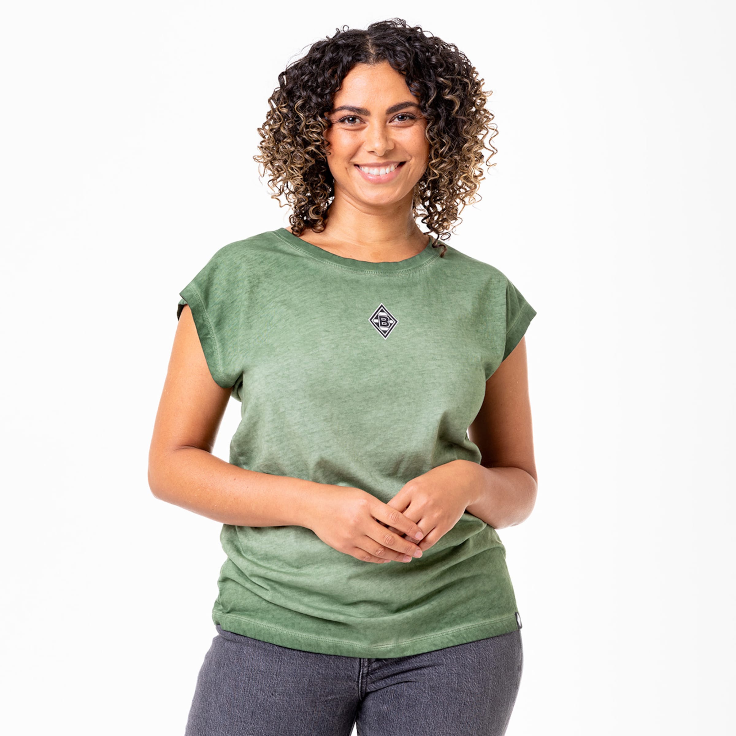 Frau mit lockigen Haaren trägt ein grünes T-Shirt mit einem kleinen Logo vorne.