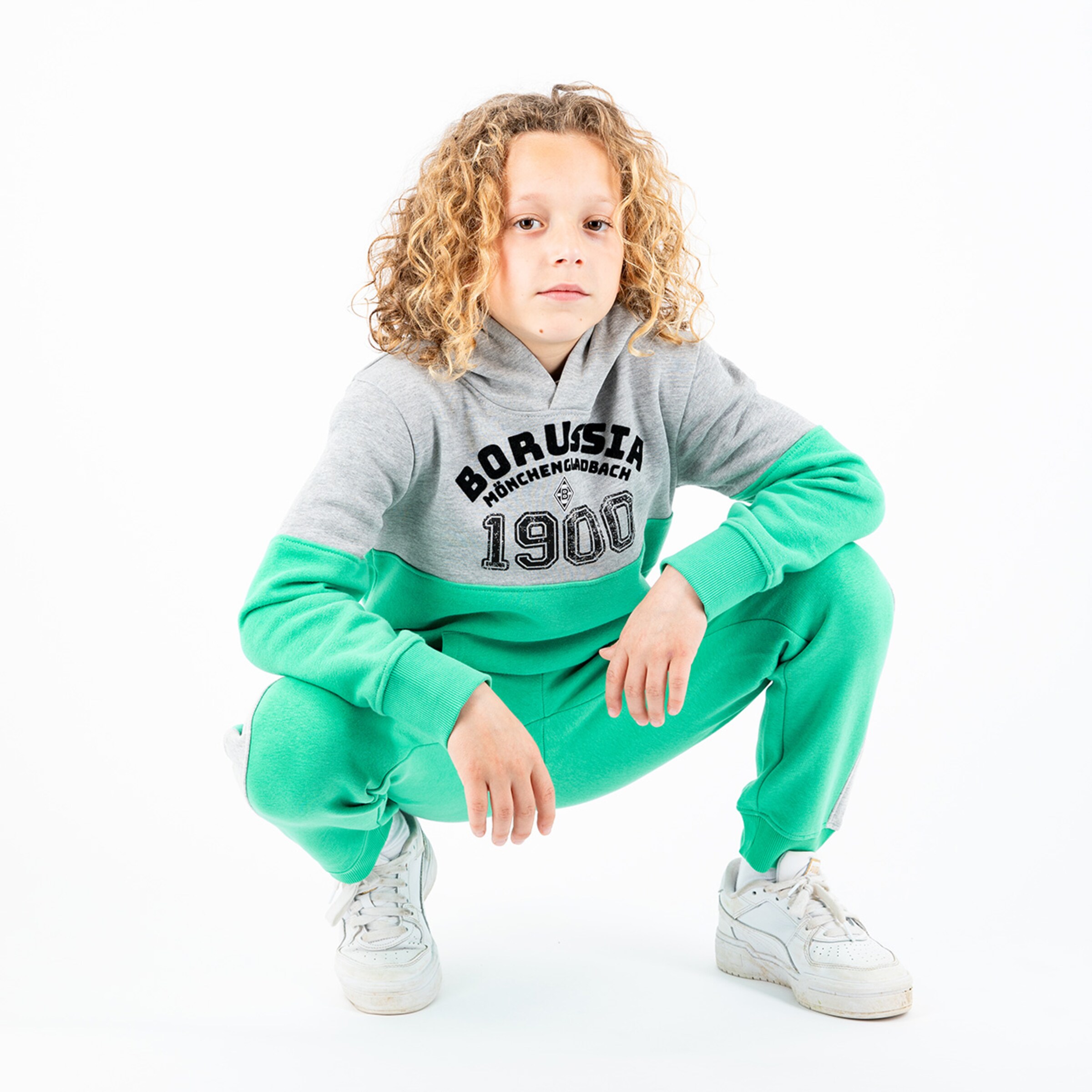 Ein Junge mit lockigen Haaren in einem grauen und grünen Jogginganzug von Borussia Mönchengladbach der in einer knienden Position posiert.
