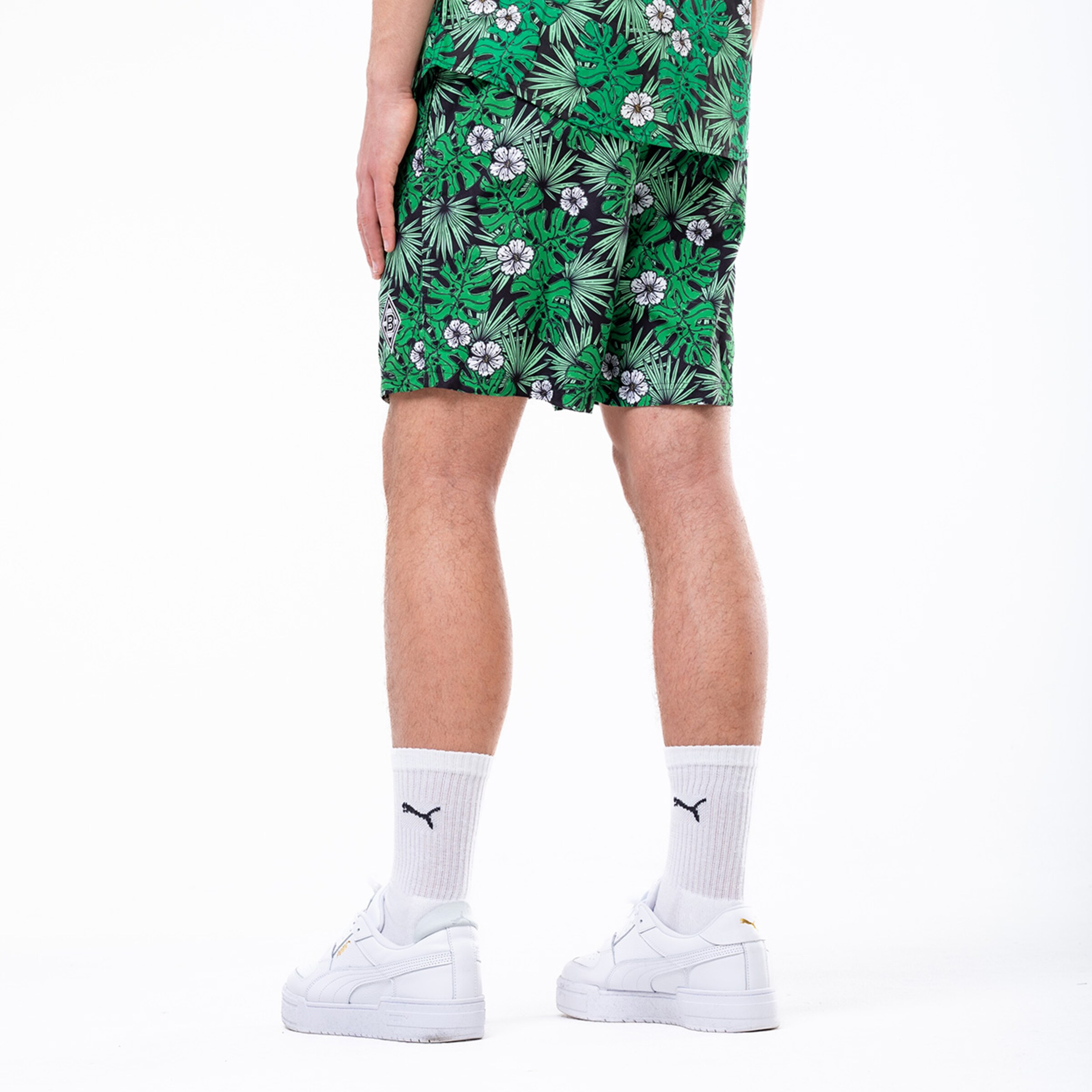 Rückansicht einer Person in grünen Shorts mit Tropenmuster, weißen Puma-Socken und weißen Sneakern vor weißem Hintergrund.