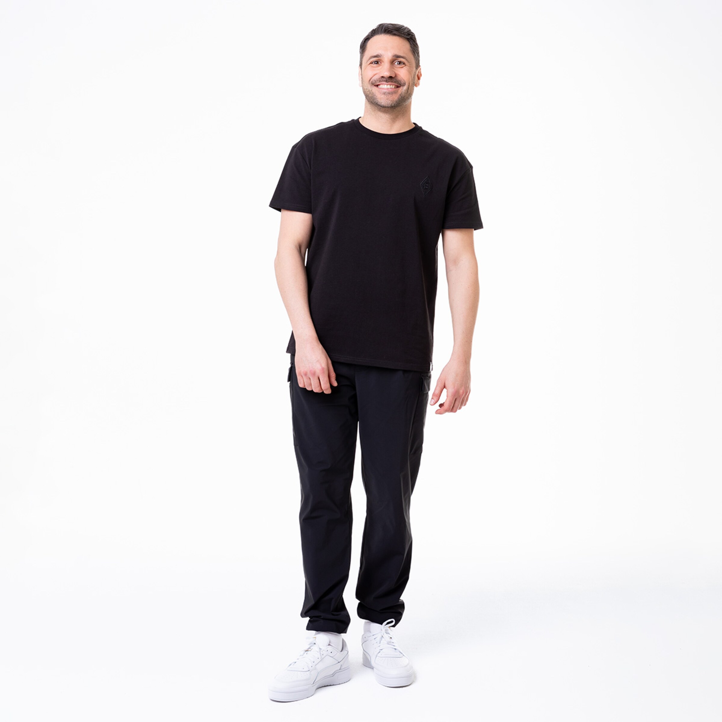 Mann lächelnd in schwarzem T-Shirt und schwarzen Jogginghosen steht aufrecht mit weißen Sneakers.