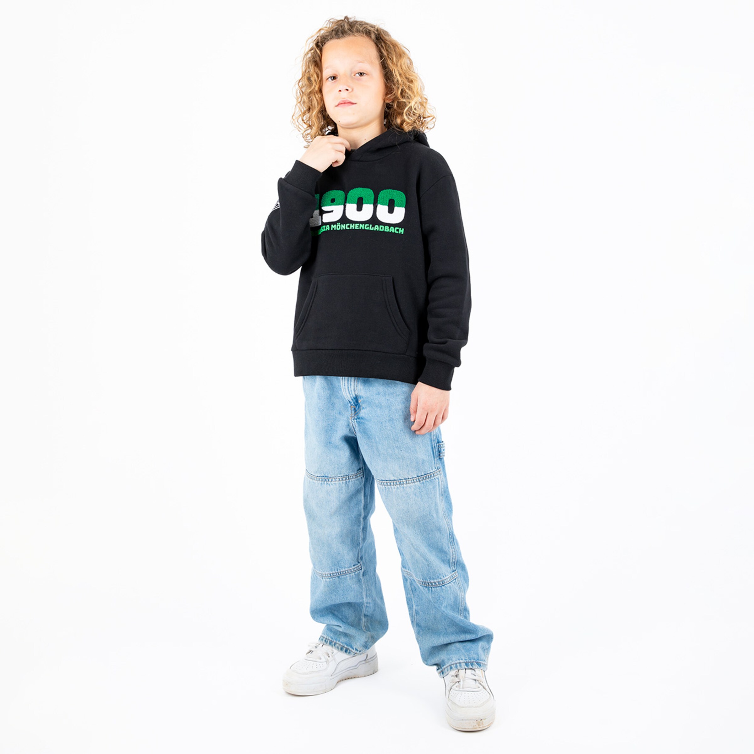 A boy with curly hair wearing a black hoodie with green lettering and blue jeans.