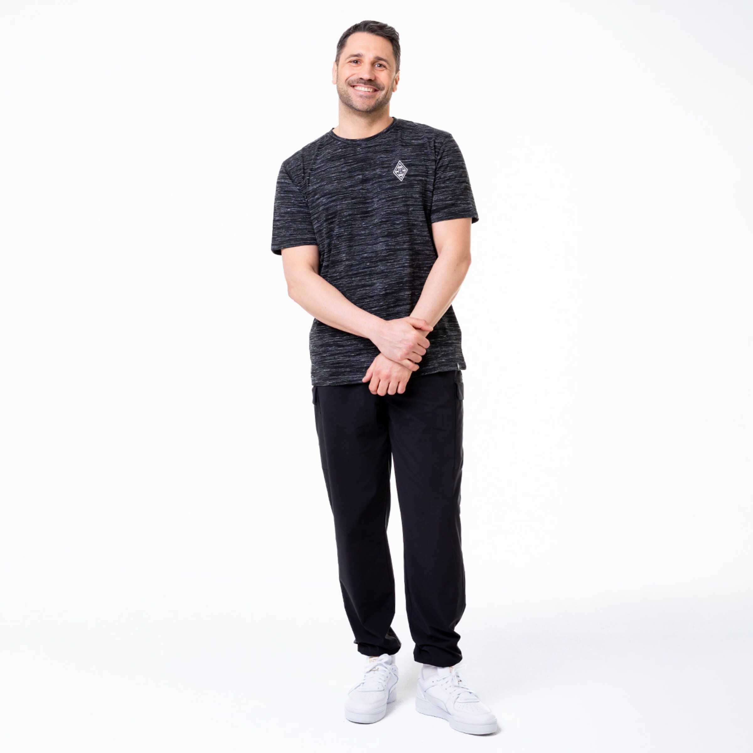 Ein Mann steht lächelnd in sportlicher Kleidung schwarze Shorts und T-Shirt.