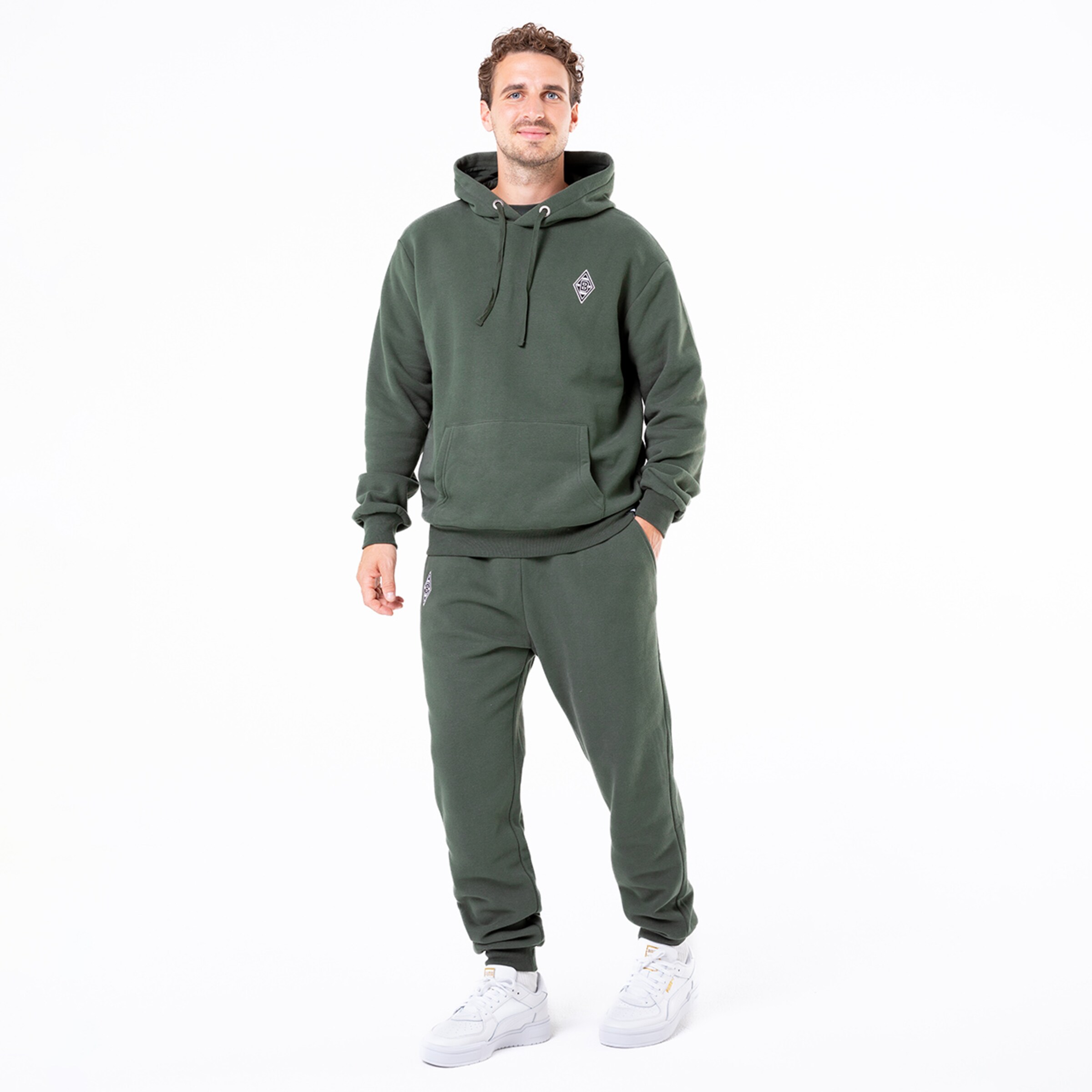 Mann in grünem Kapuzenpullover und Jogginghose steht lächelnd mit einer Hand in der Hose.