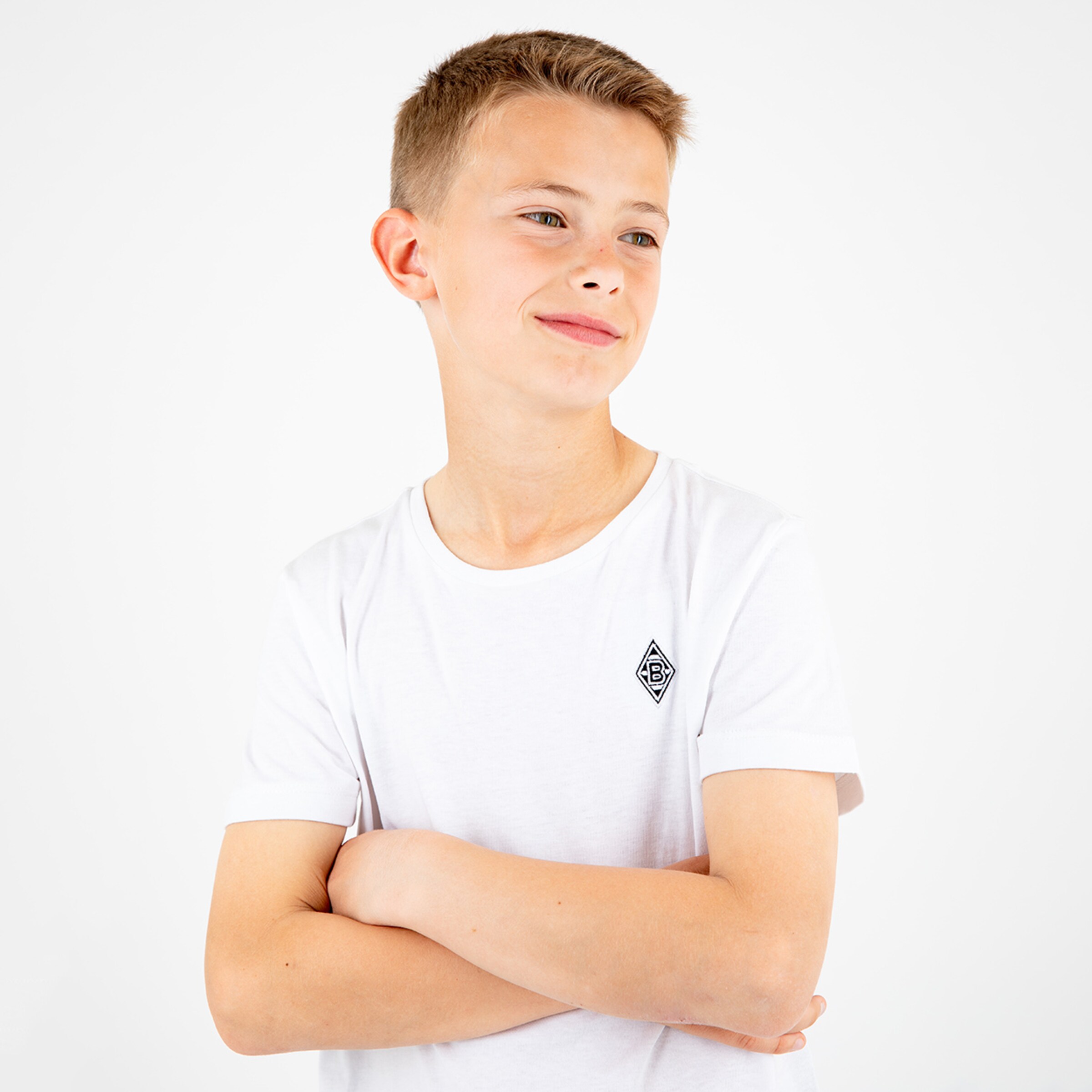 Ein Junge mit kurzen blonden Haaren trägt ein weißes T-Shirt und hat die Arme vor der Brust verschränkt.