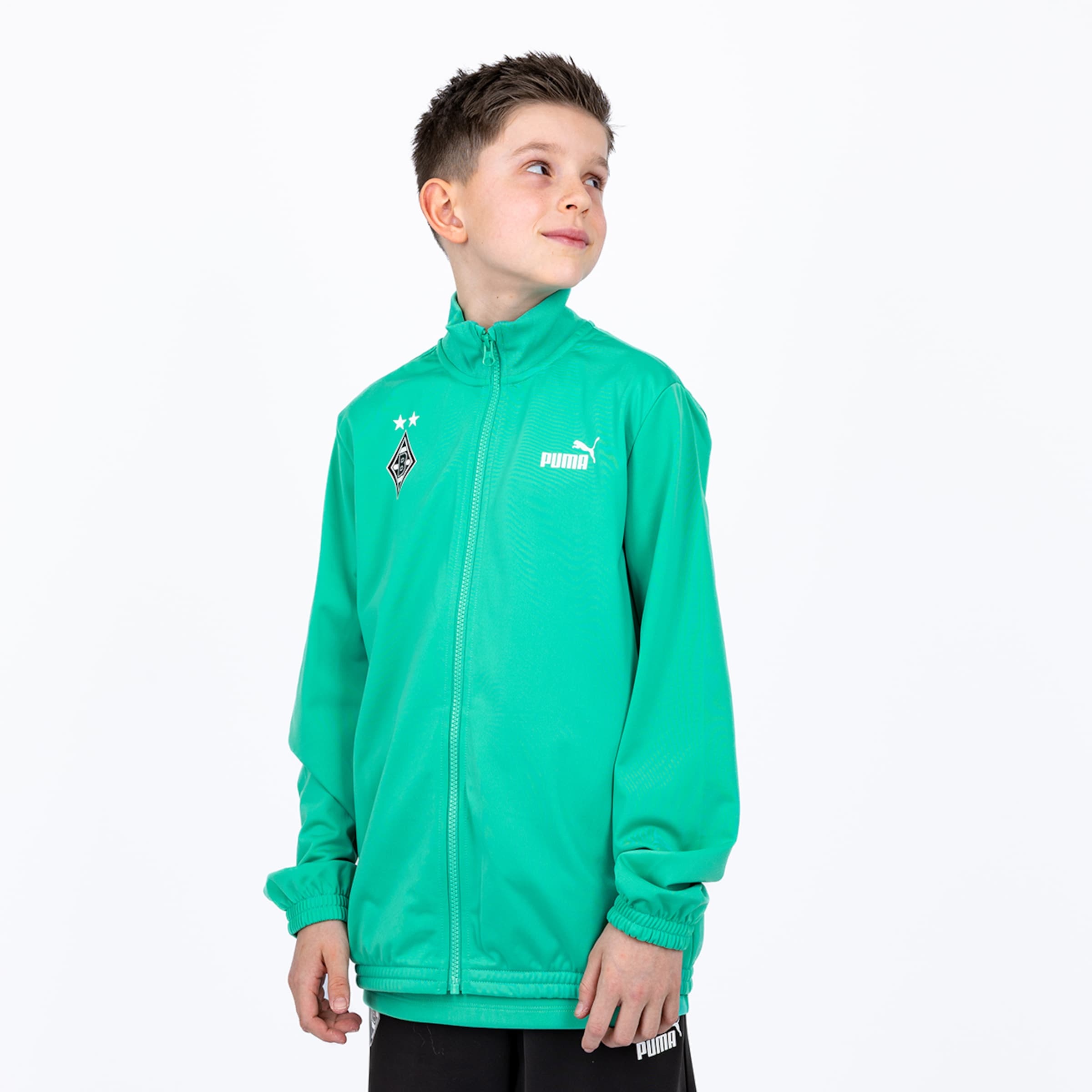 A boy wearing a green Puma Borussia Mönchengladbach track jacket and black shorts against a white background.