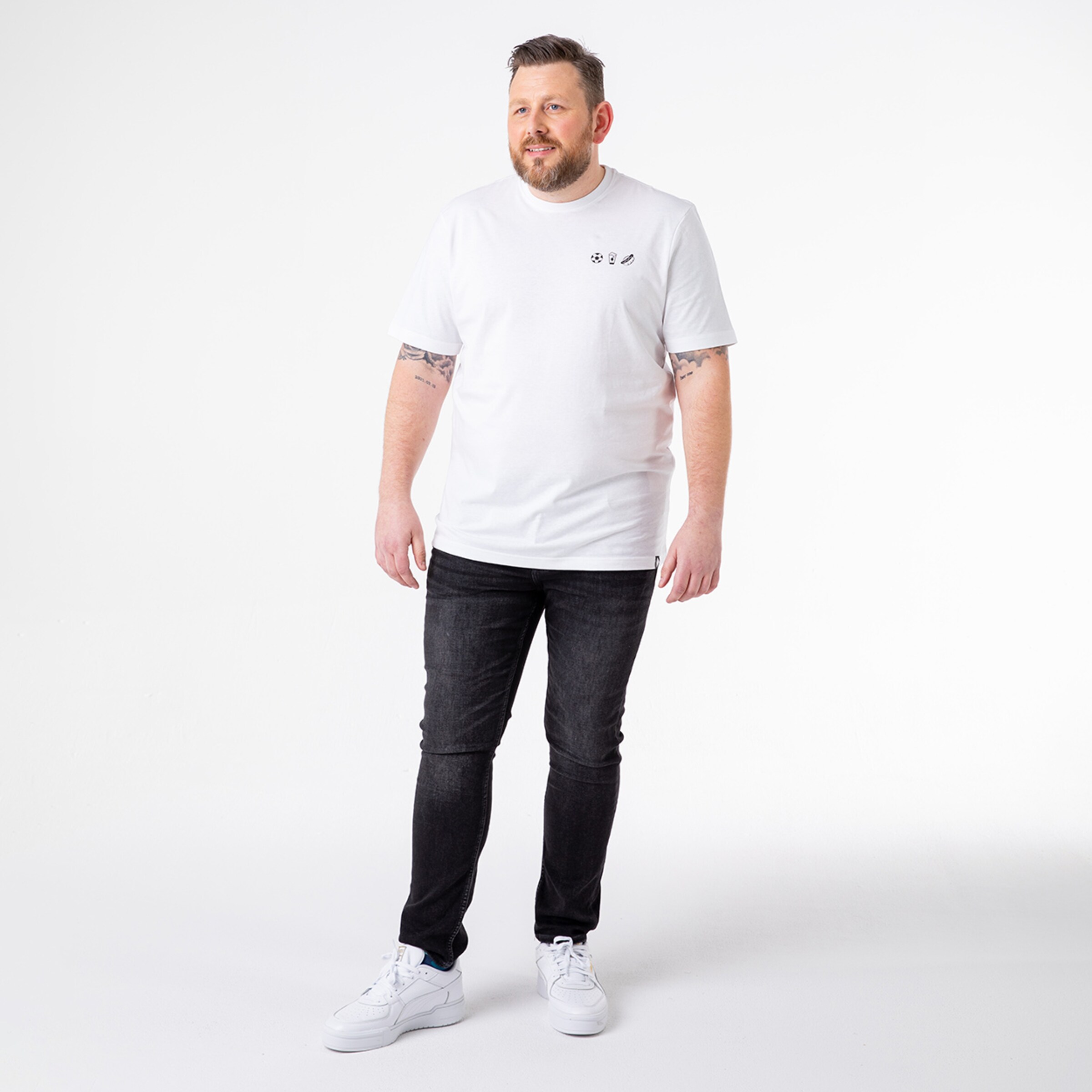 Ein Mann steht in einem weißen T-Shirt und dunklen Jeans trägt weiße Sneaker.