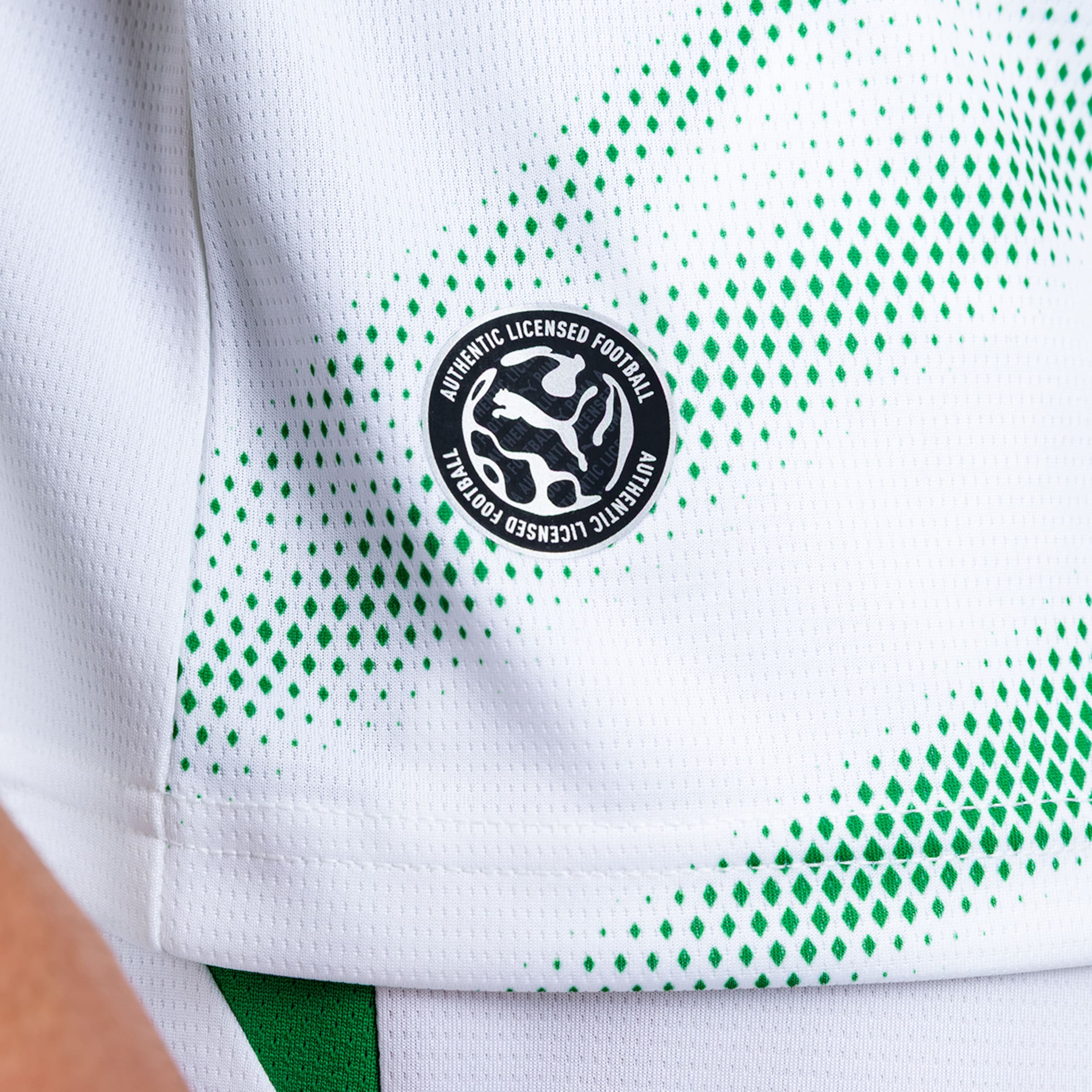 Nahaufnahme eines weißen Fußballtrikots mit grünem Rautenmuster und rundem schwarzen Puma „Authentic Licensed Football“ Patch.