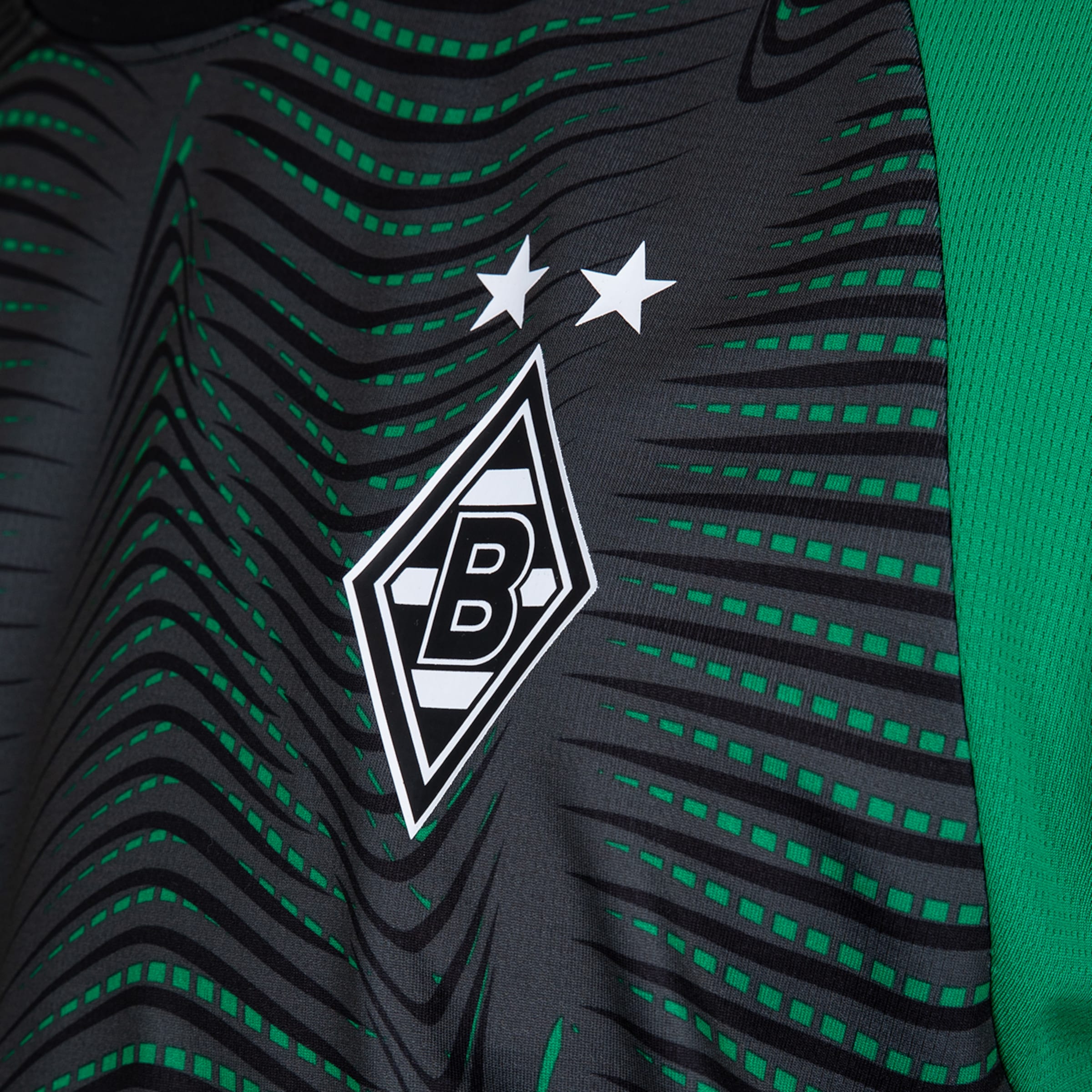 Nahaufnahme eines Borussia Mönchengladbach Fußballtrikots in Dunkelgrau und Grün. Es zeigt ein wellenförmiges Muster mit schwarzen Linien und grünen Quadraten, das Logo mit 'B' und zwei Sternen, sowie einen grünen Netzärmel.