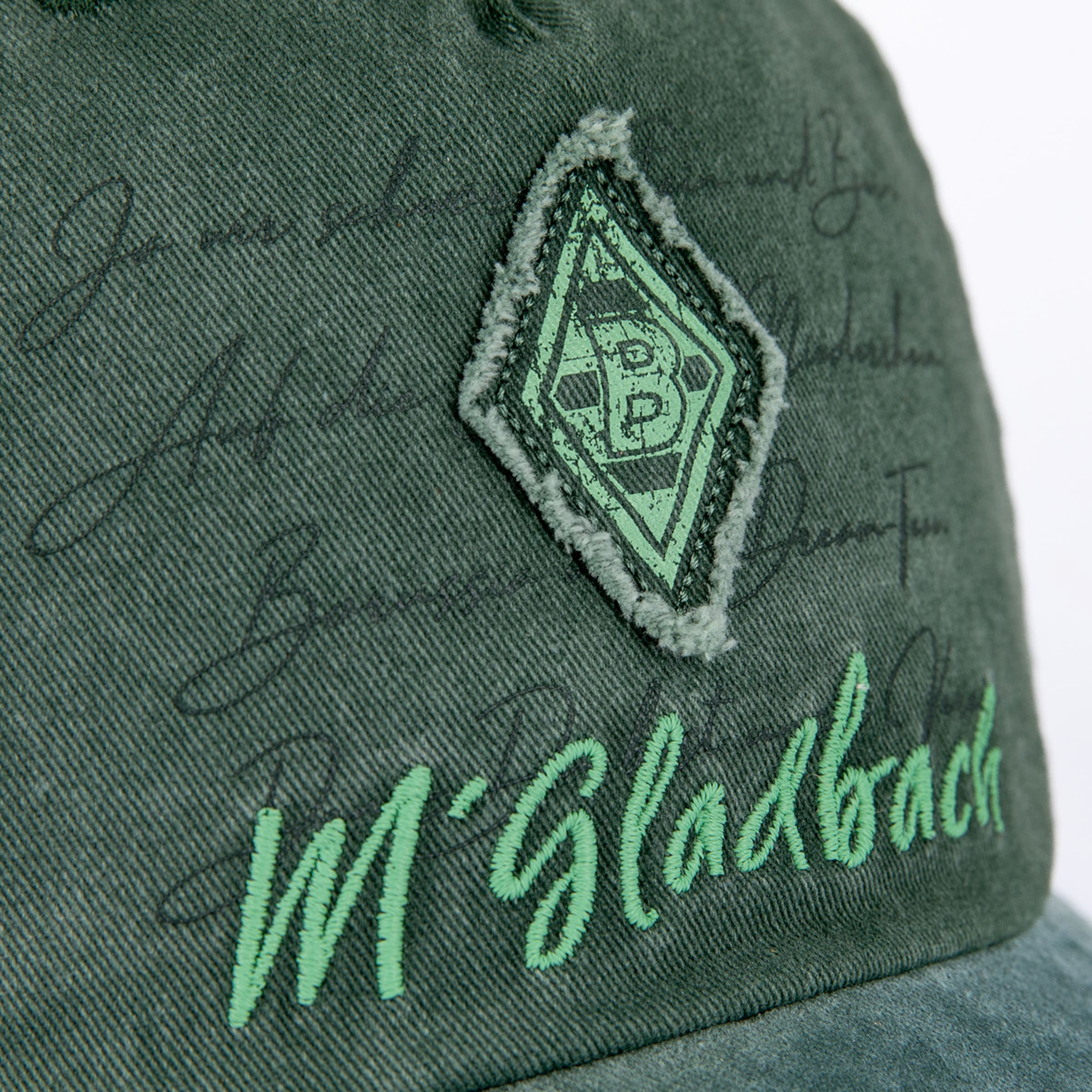Nahaufnahme einer grünen Gladbach-Cap mit Rauten-Logo und hellgrünem „M'gladbach“-Schriftzug. Dezenter Print im Hintergrund.