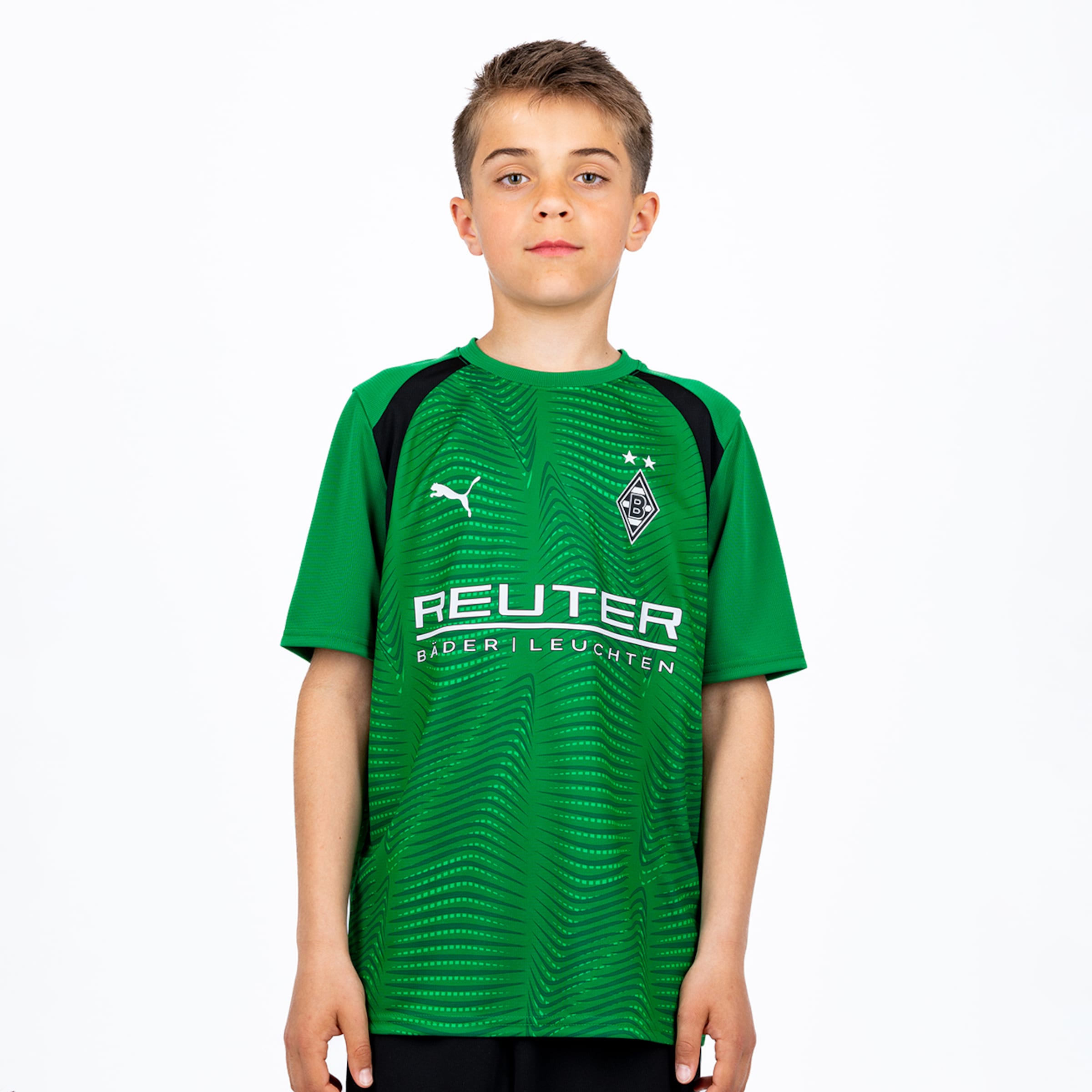 Ein junger Junge trägt ein grünes Borussia Mönchengladbach Fußballtrikot mit schwarzen Schultern, einem Puma-Logo, dem Vereinswappen mit zwei Sternen und dem Sponsor „REUTER BÄDER | LEUCHTEN“. Er steht vor einem weißen Hintergrund.