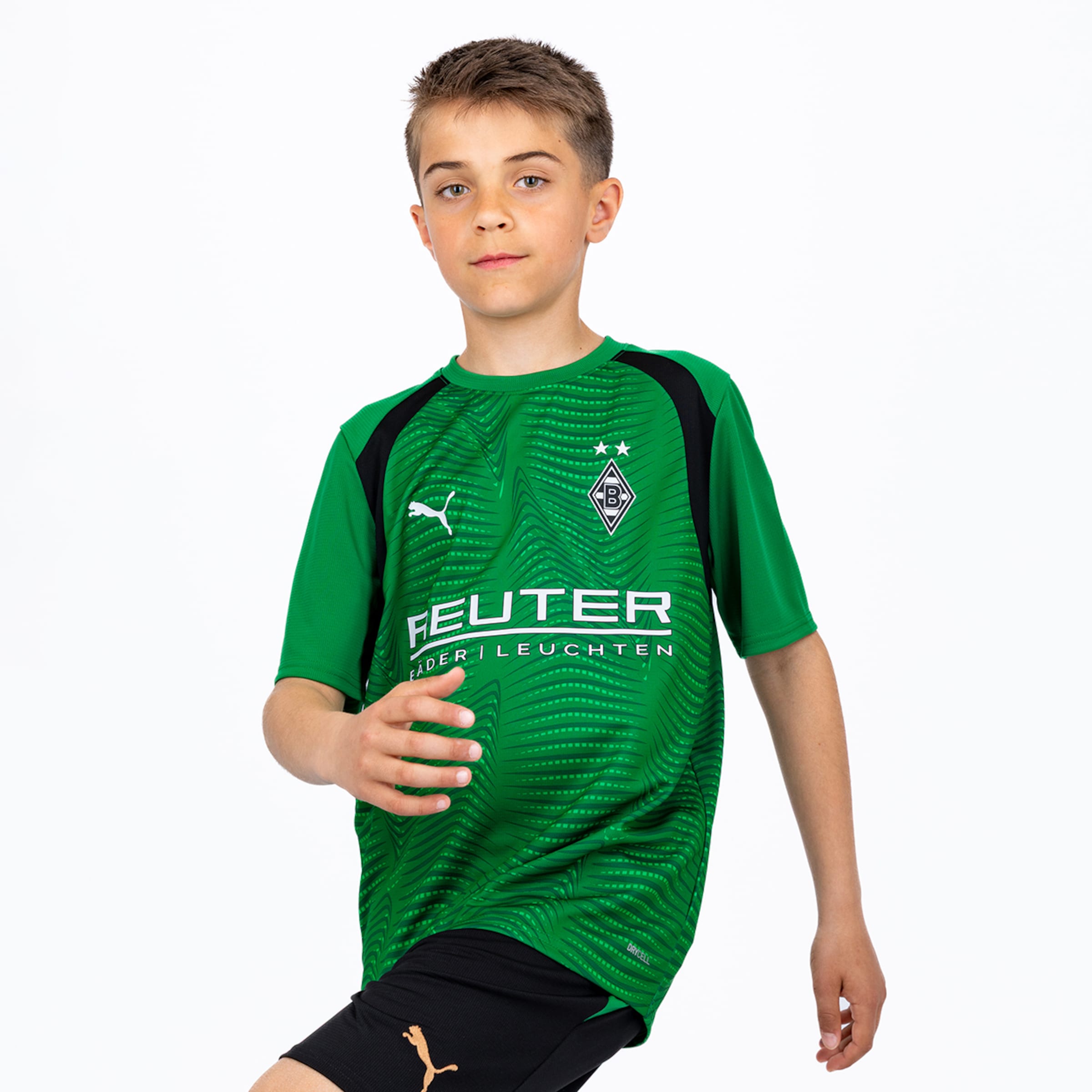 Ein junger Junge trägt ein grünes Fußballtrikot mit schwarzen Schulterpartien und schwarze Shorts. Er posiert vor weißem Hintergrund. Das Trikot zeigt das Puma-Logo, ein Vereinswappen mit zwei Sternen und den Sponsor 'FEUTER FÄDER LEUCHTEN'.