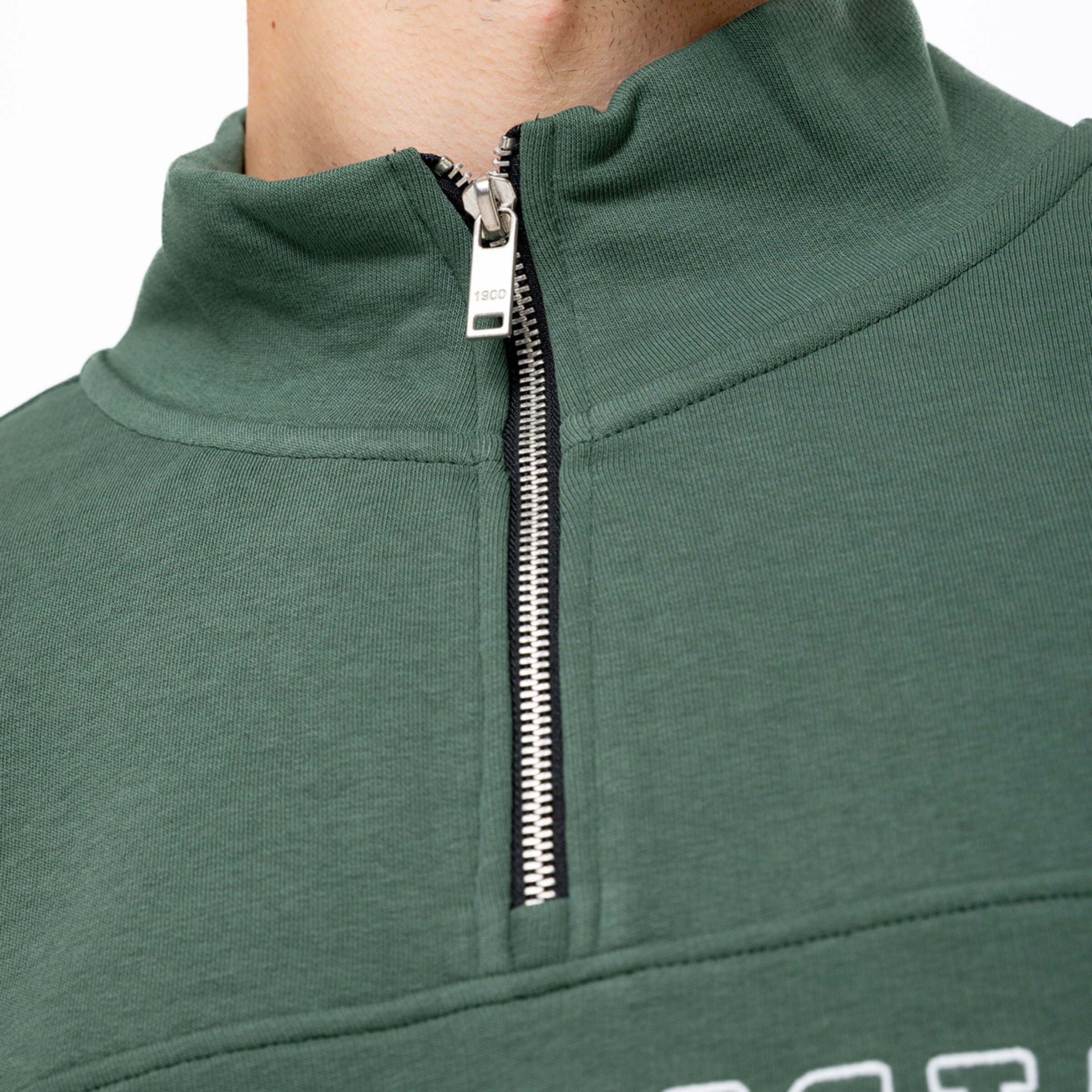 Nahaufnahme eines dunkelgrünen Troyers mit silbernem Reißverschluss und hohem Kragen. Auf dem Reißverschlussgriff steht die Zahl 1900.