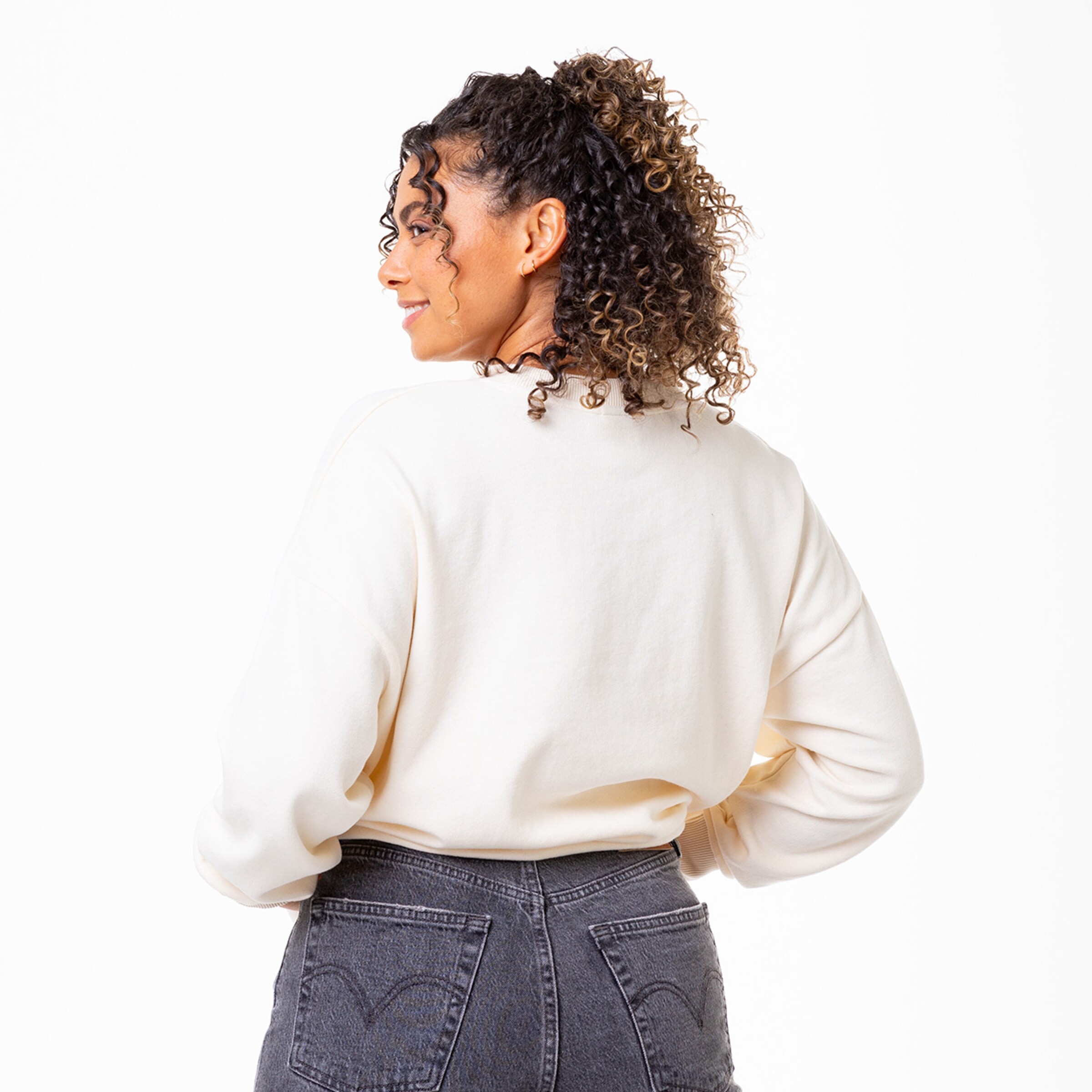 Eine Frau mit lockigem Haar schaut über ihre Schulter. Sie trägt einen hellen Sweater und hat eine lockere Jeans an.