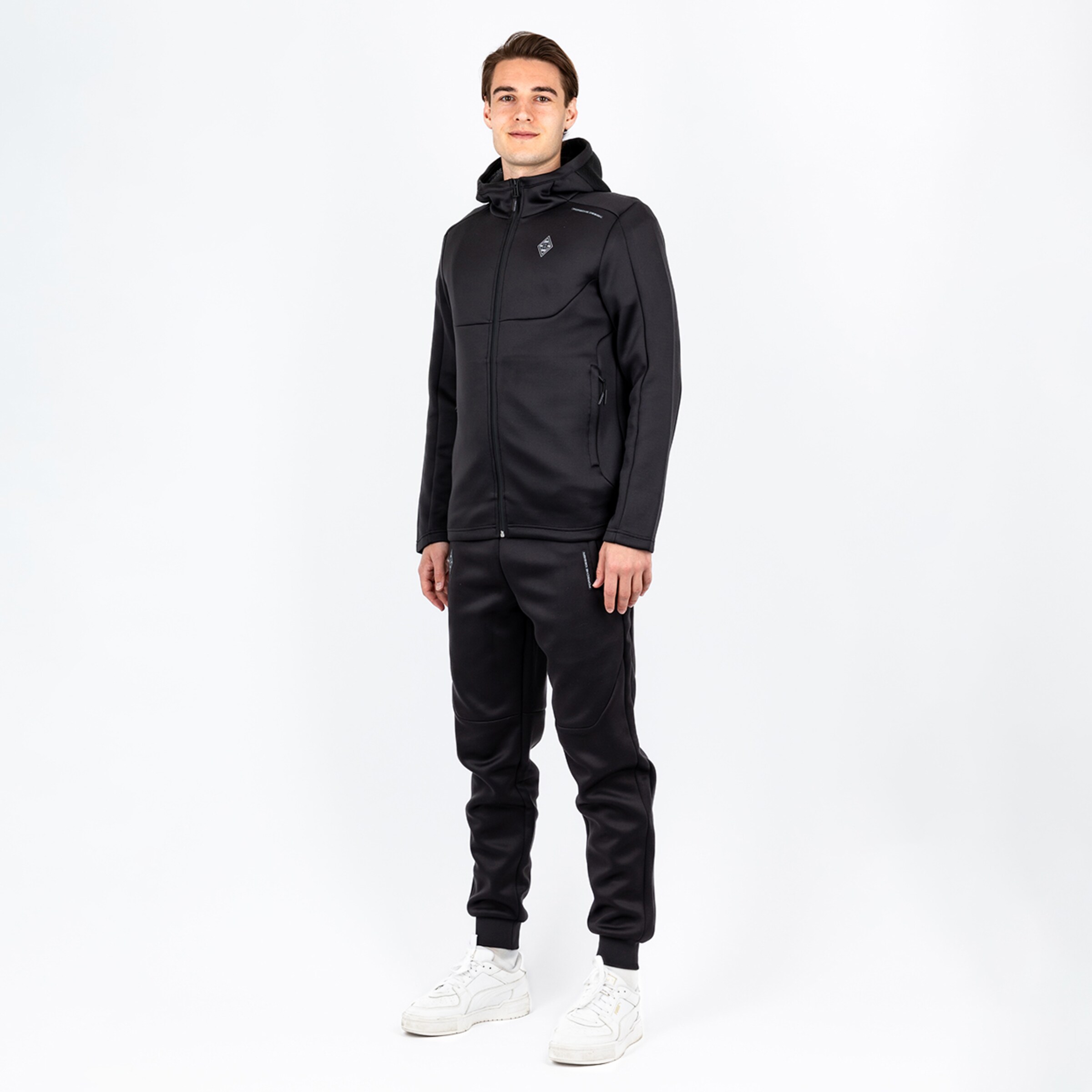 Mann in schwarzem Sportanzug mit Kapuze weißen Sneakers und entspanntem Gesichtsausdruck.