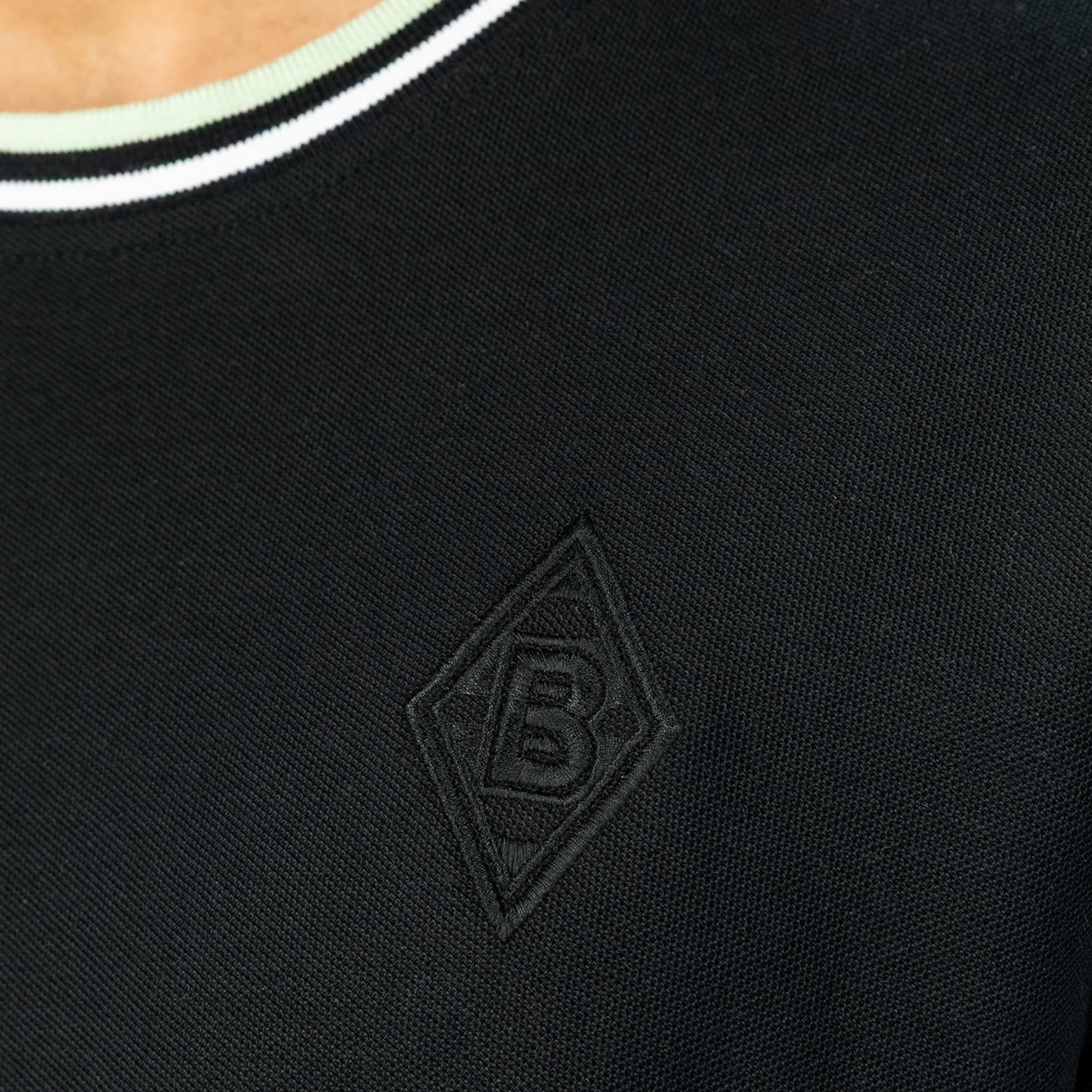 Nahaufnahme eines schwarzen Oberteils mit dezentem, schwarz gesticktem Rauten-Logo (B) und weiß-hellgrün gestreiftem Kragen.