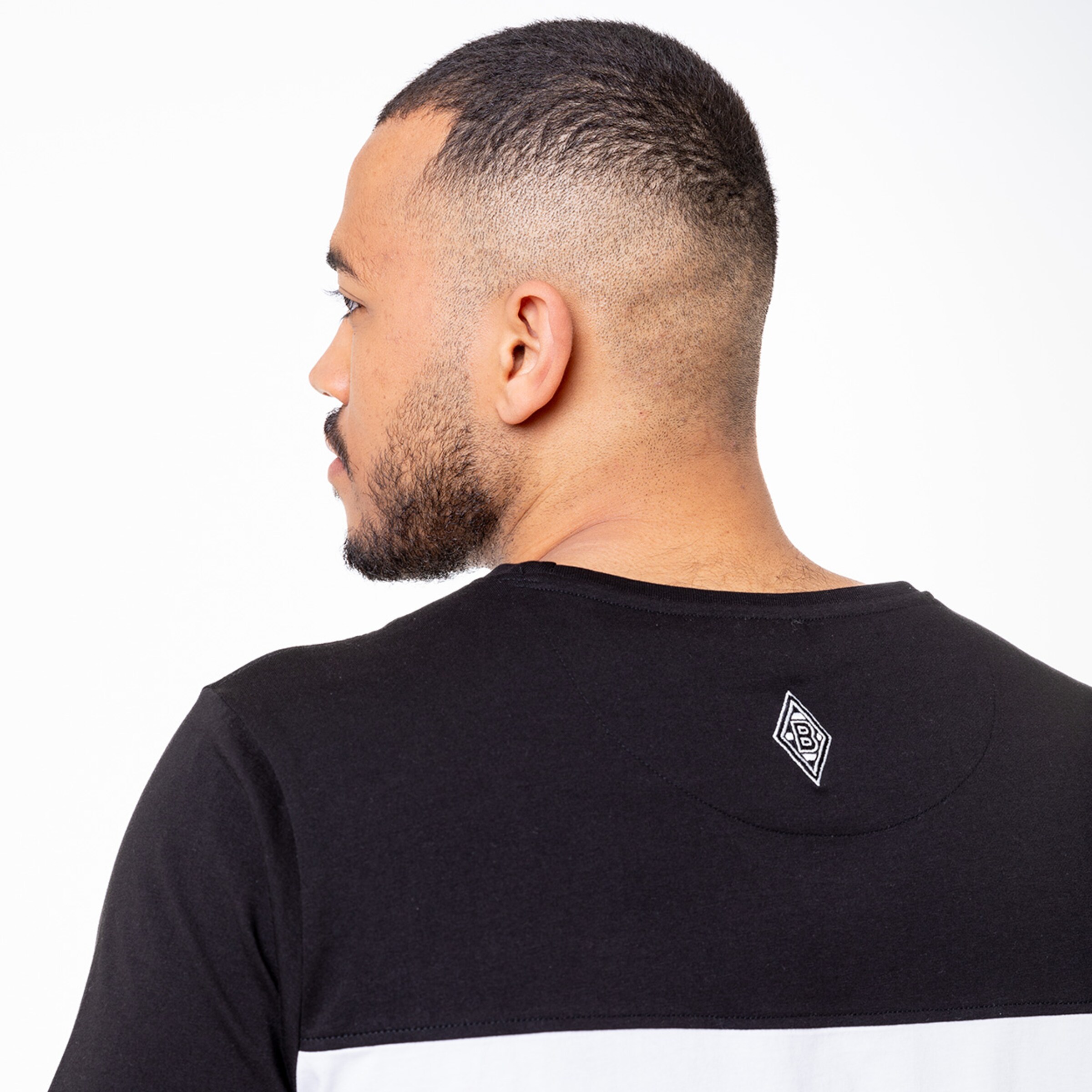 Ein Mann mit kurzen Haaren und einem Bart der ein schwarzes T-Shirt mit einer weißen und grauen Farbblockierung trägt. Das T-Shirt hat ein kleines Logo auf dem Rücken.