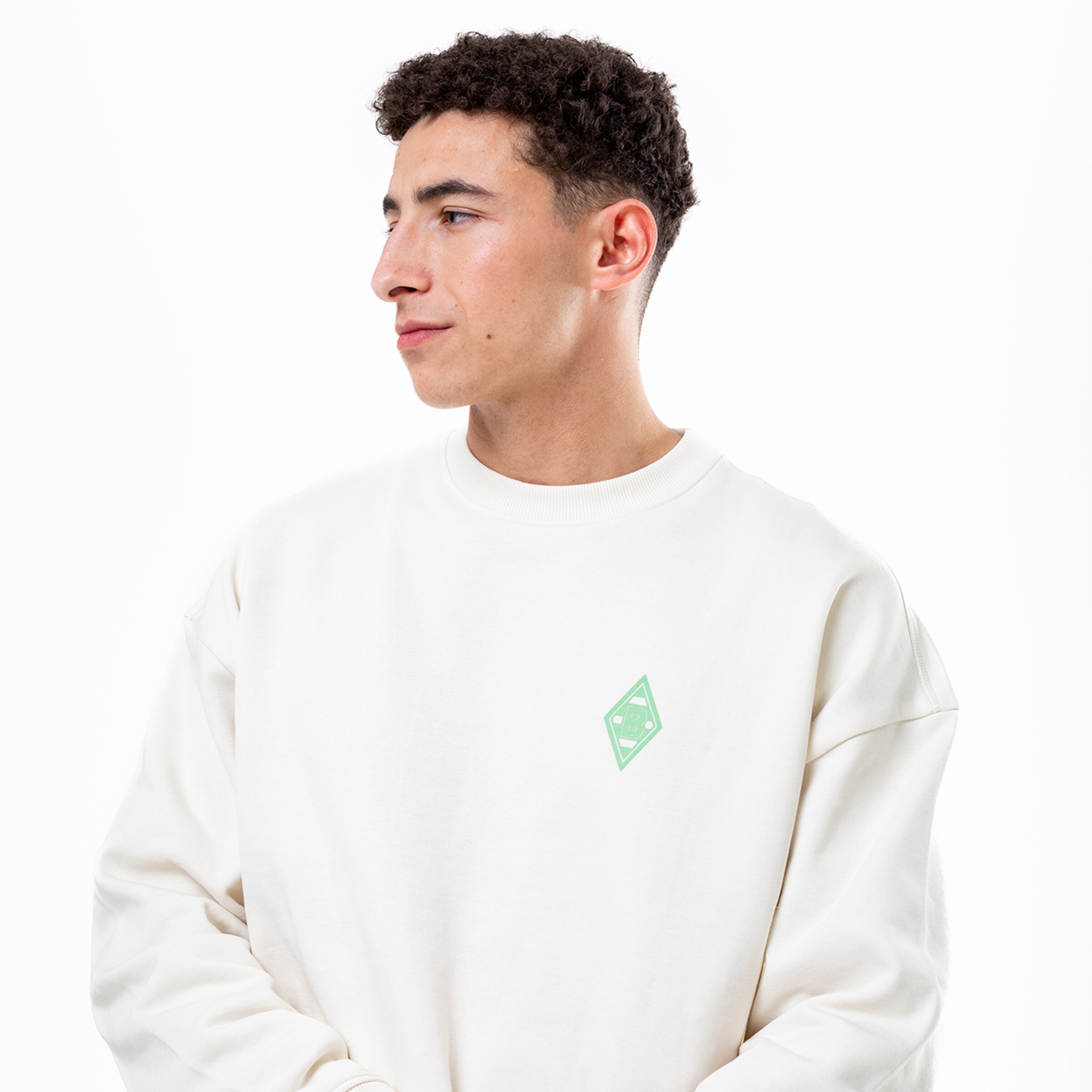 Junger Mann mit lockigem Haar trägt einen weißen Sweatshirt mit grünem Logo.