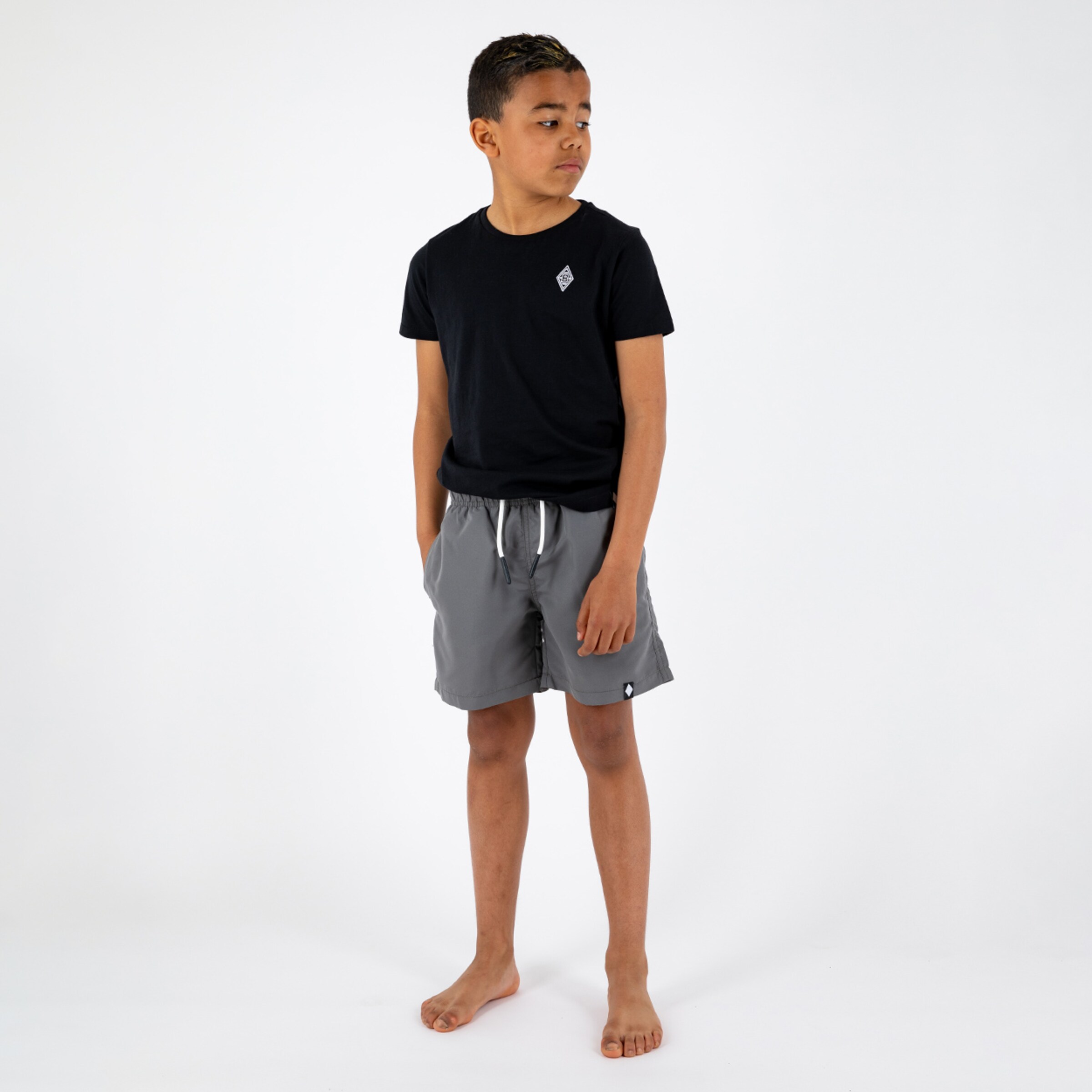 Ein Junge steht mit einer dunklen T-Shirt und grauen Shorts barfuß mit den Händen in den Taschen.