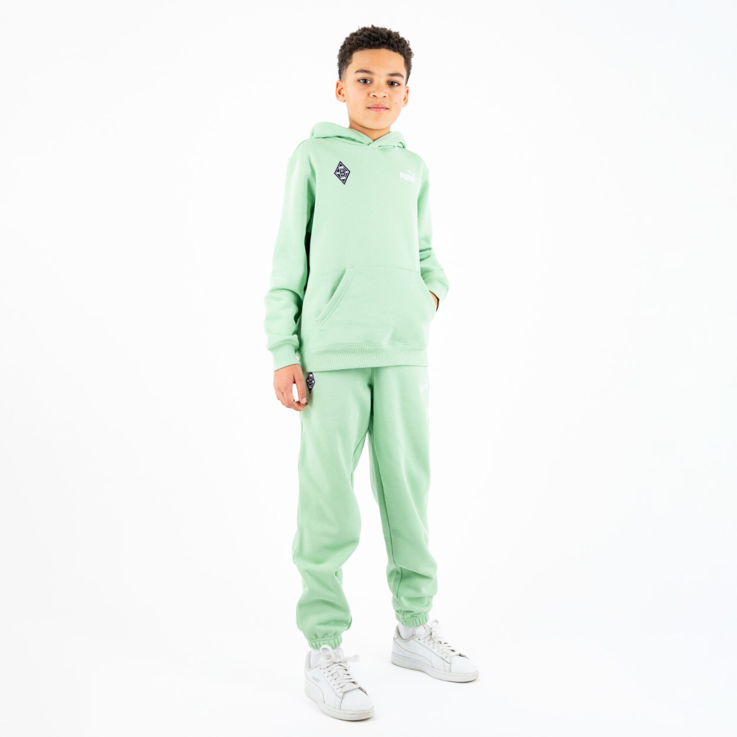 Ein Junge in einer mintgrünen Jogginganzug mit Kapuze und weißen Sneakers steht in aufrechter Position.