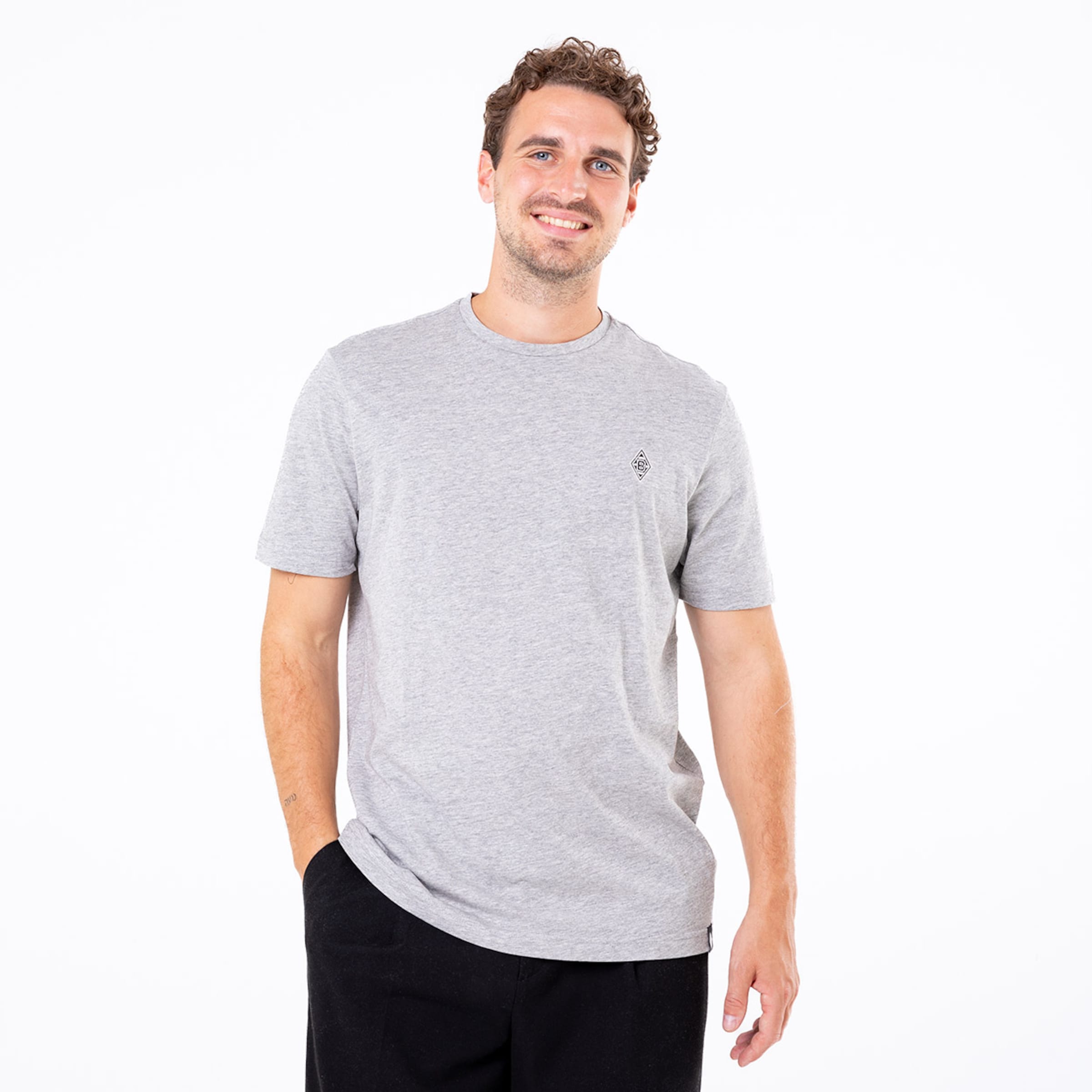 Mann in grauem T-Shirt mit kurzer Ärmel und schwarzer Hose lächelt und hat eine Hand in der Hosentasche.