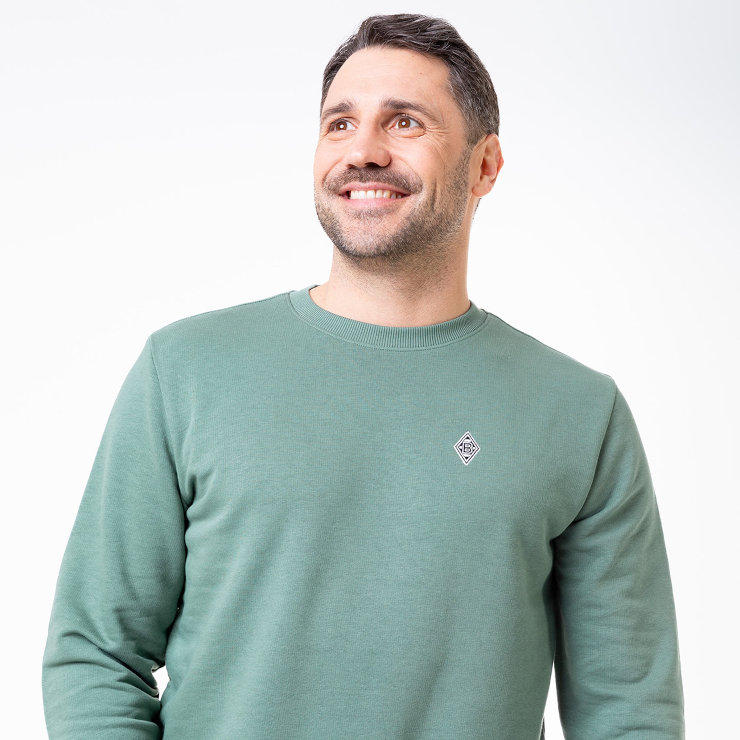 Ein Mann mit einem freundlichen Lächeln trägt einen grünen Pullover mit einem kleinen Logo.