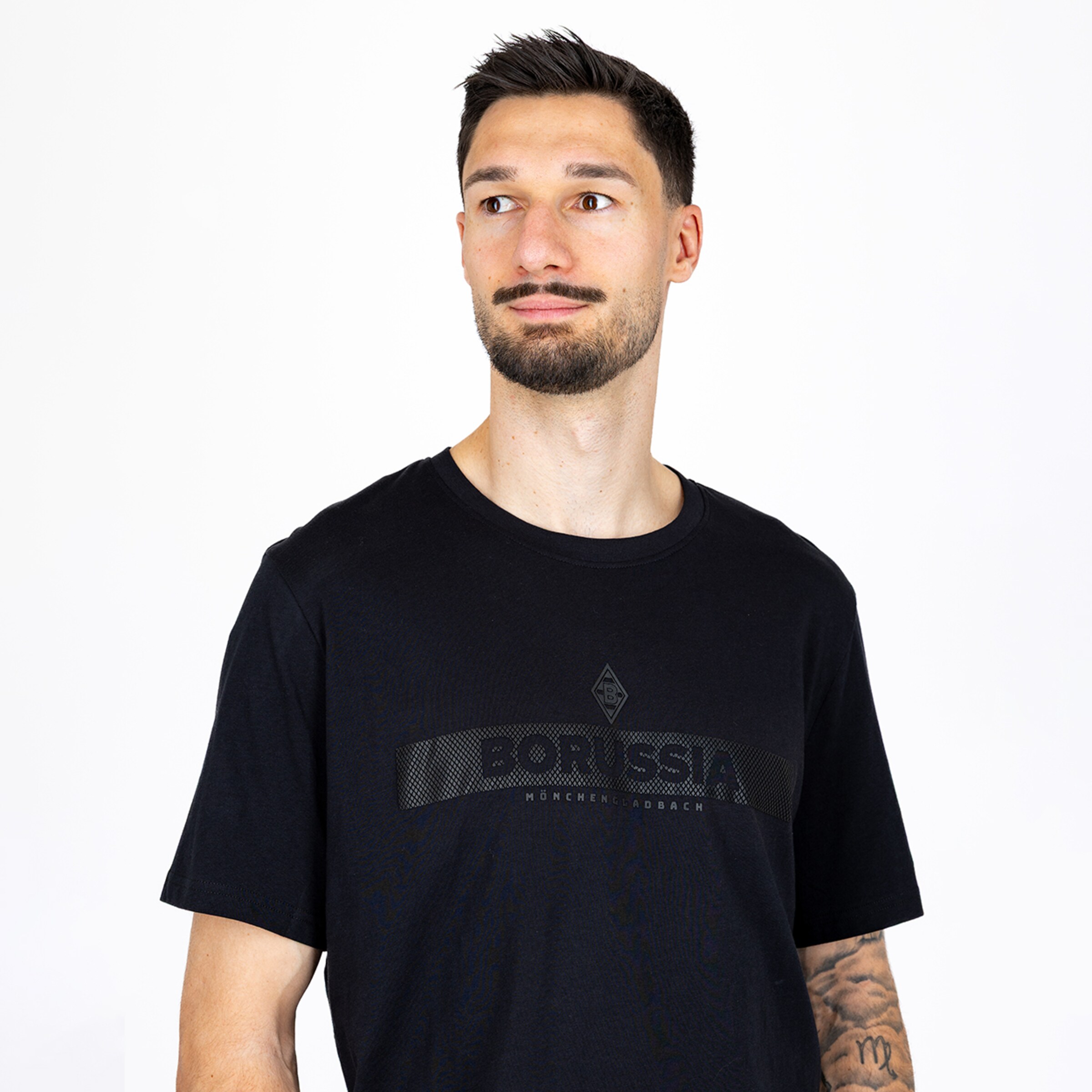 Junger bärtiger Mann in schwarzem T-Shirt mit Borussia Mönchengladbach-Schriftzug vor weißem Hintergrund.