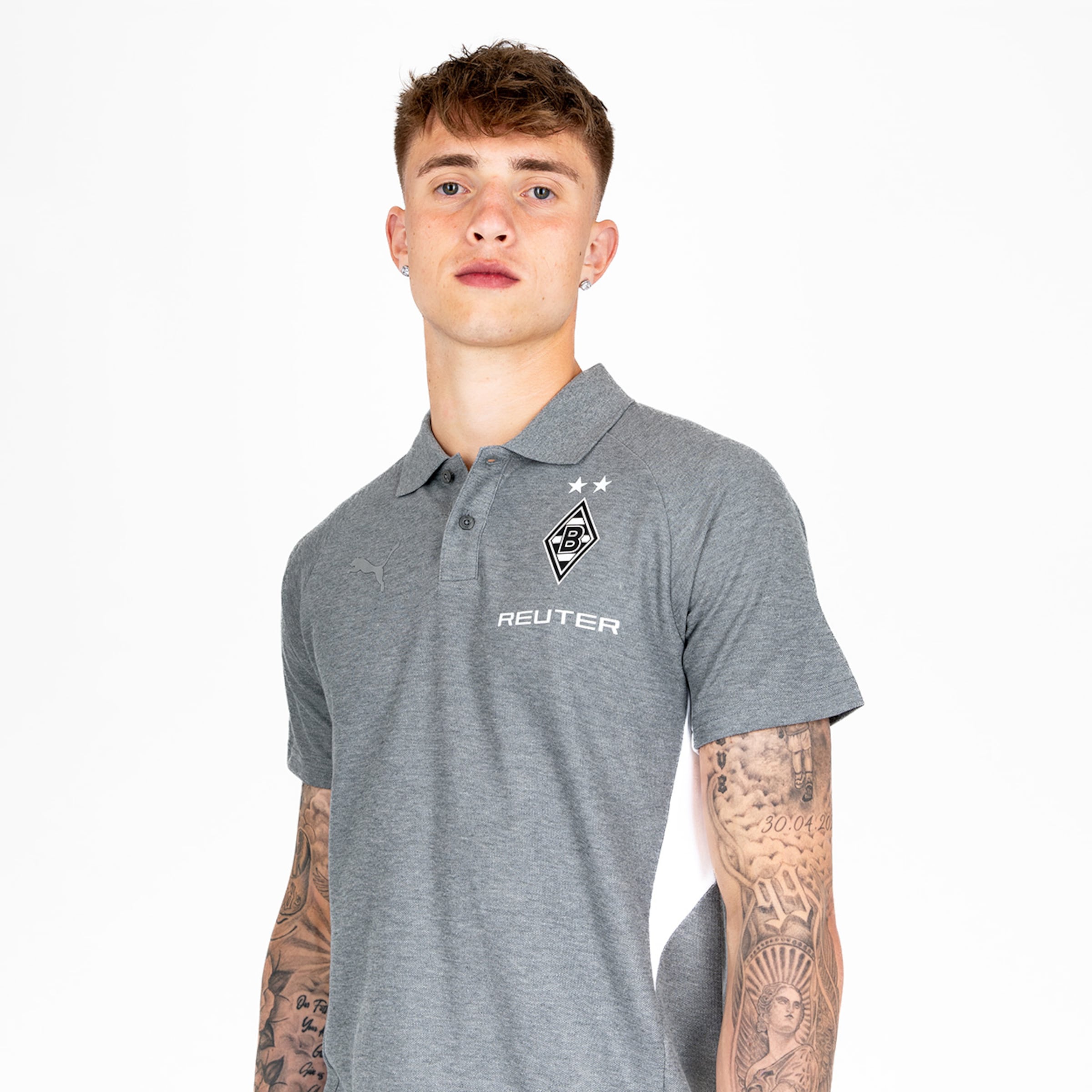 Ein Mann mit tätowierten Armen trägt ein graues Poloshirt mit dem Borussia Mönchengladbach Logo und der Aufschrift REUTER.