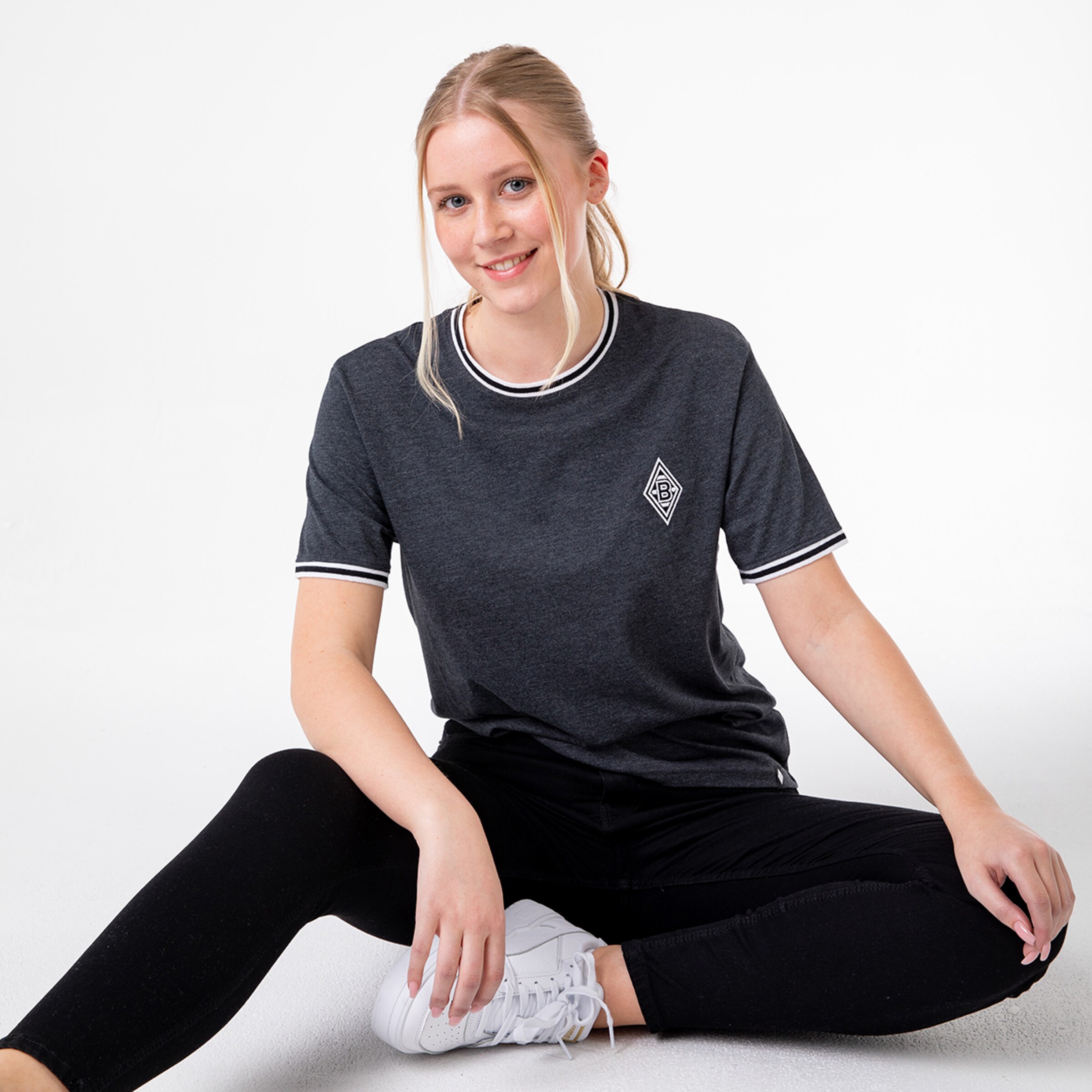 Eine Frau sitzt auf dem Boden trägt ein schwarzes T-Shirt mit weißem Rundhalsausschnitt und hellen Streifen.