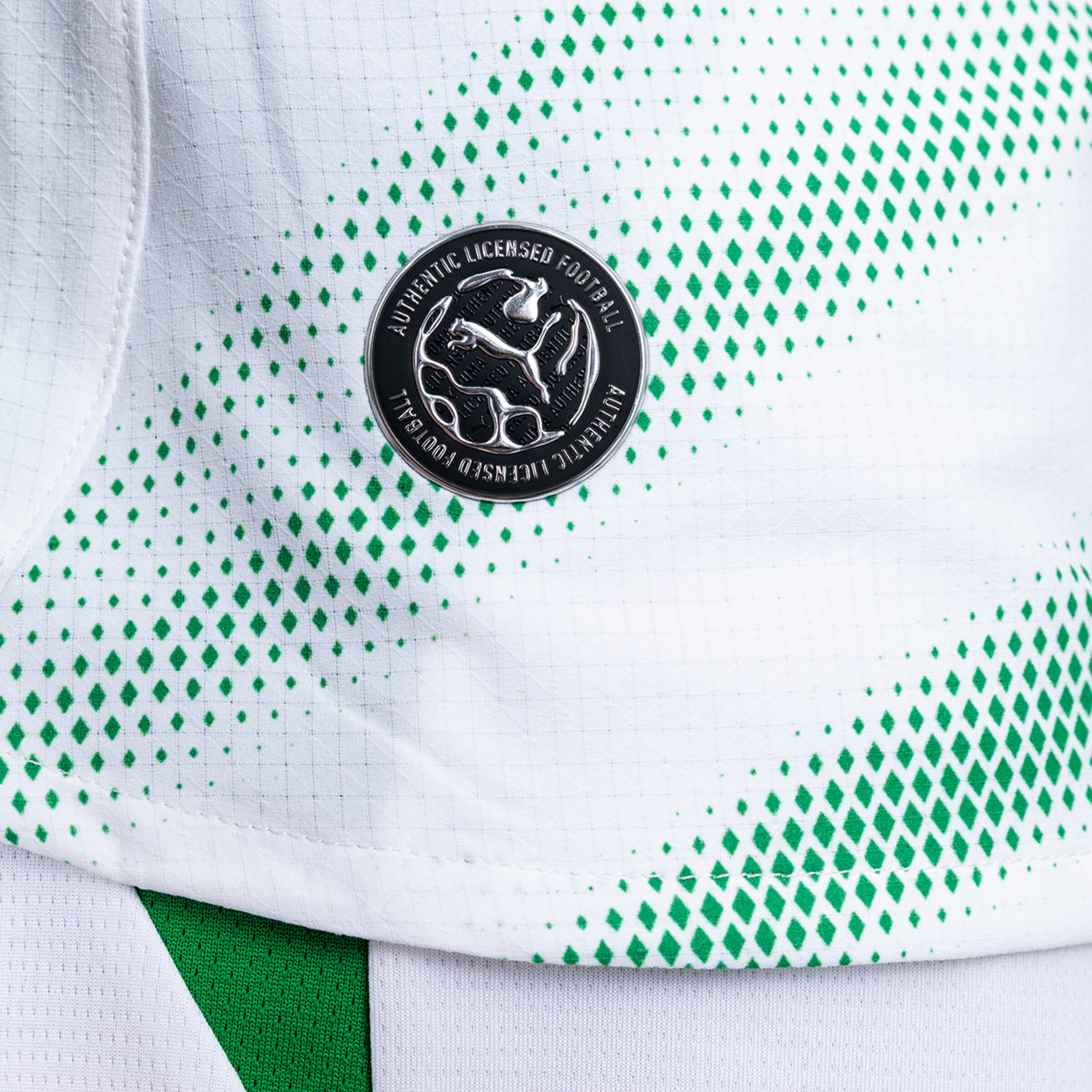 Close-up of a white jersey with green diamond patterns and a round black and silver "Authentic Licensed Football" Puma badge.