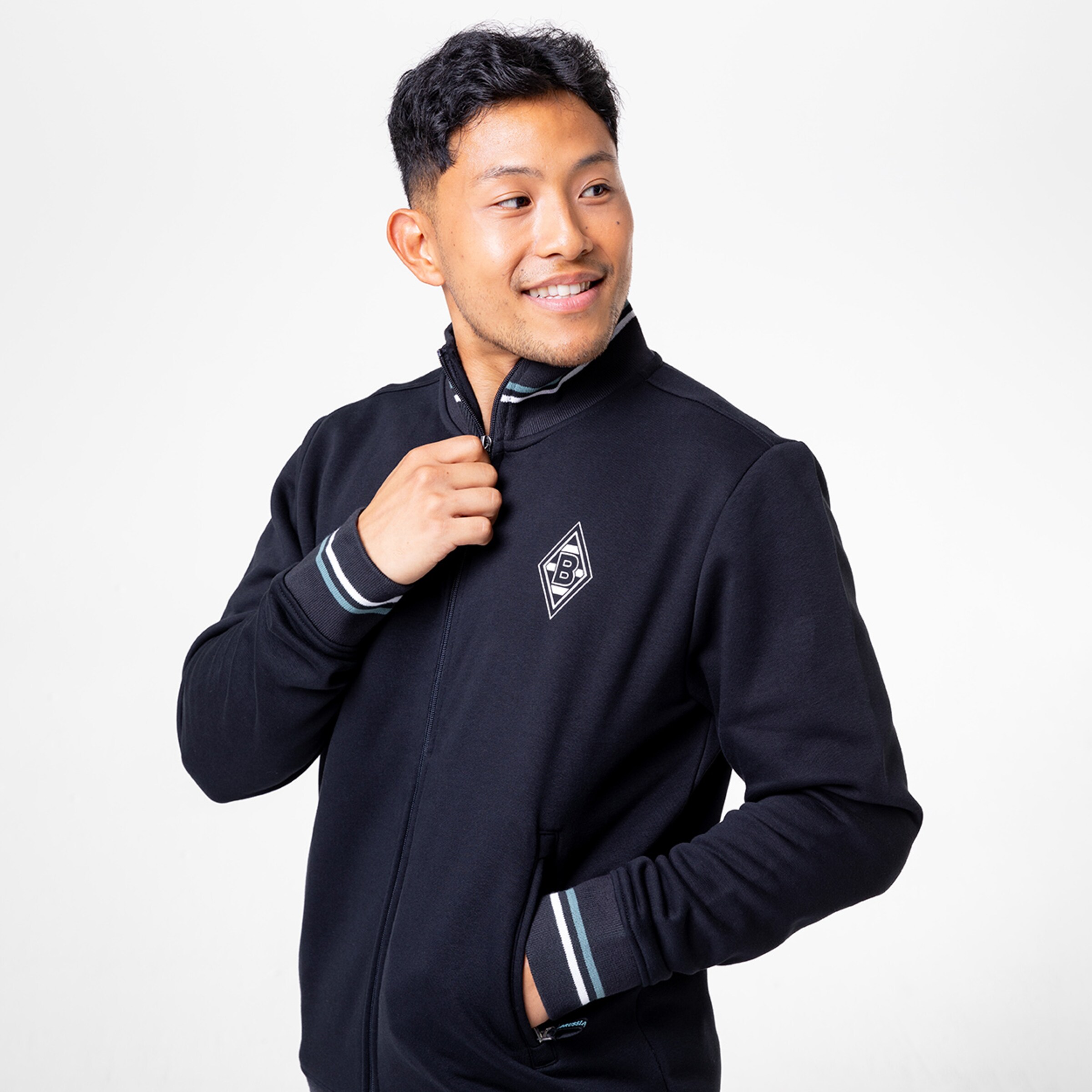 Junger Mann in schwarzem Sportjackett mit Logo lächelt und schaut zur Seite.