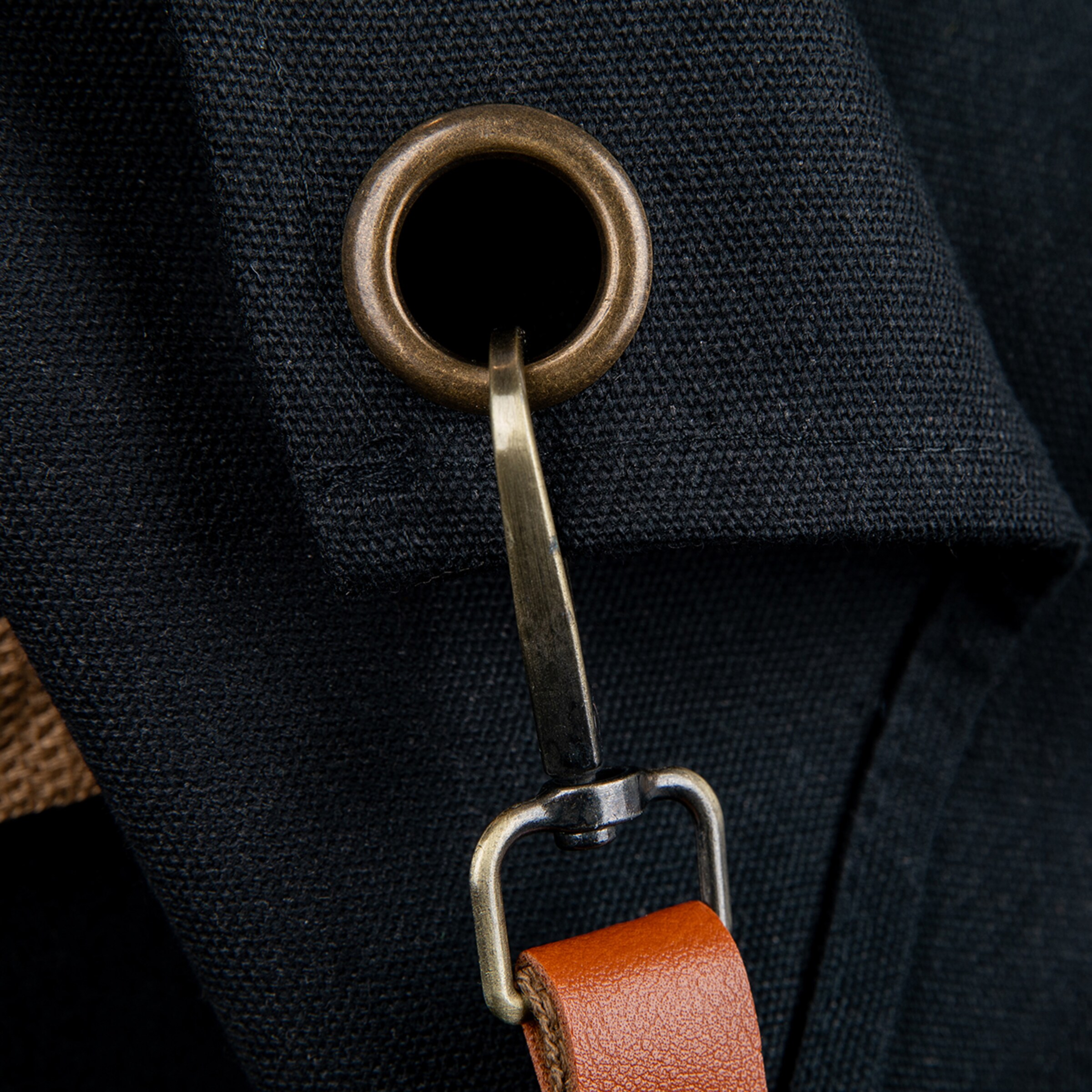 Detail shot of dark, woven fabric with a bronze eyelet, securing a metal hook to a rich brown leather strap. Emphasizes texture and robust hardware.