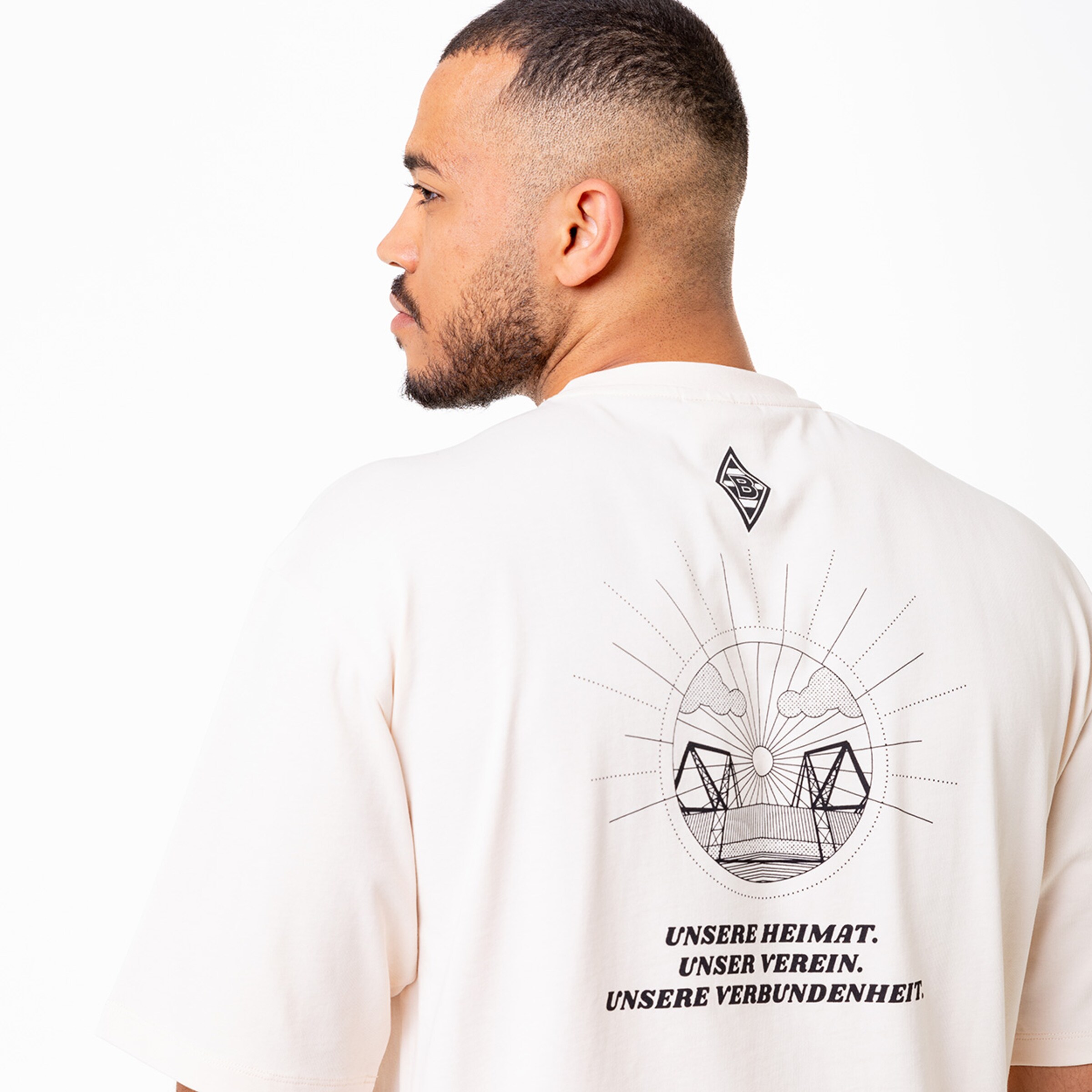 Man wearing a beige t-shirt showing a design of a stadium and the text OUR HOMELAND. OUR CLUB. OUR UNITY. on the back.