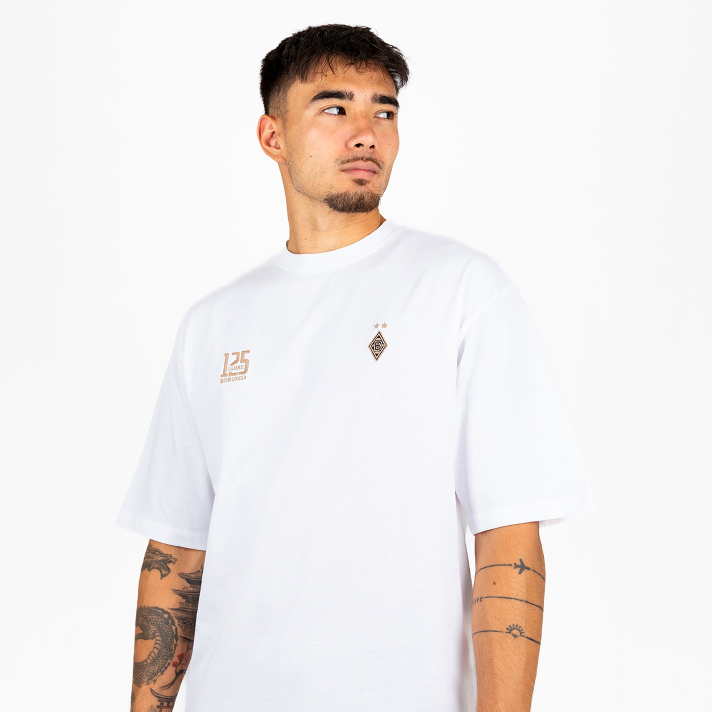 Junger Mann mit Arm-Tattoos in weißem T-Shirt mit „125 JAHRE BORUSSIA“-Print und Vereinslogo vor weißem Hintergrund.