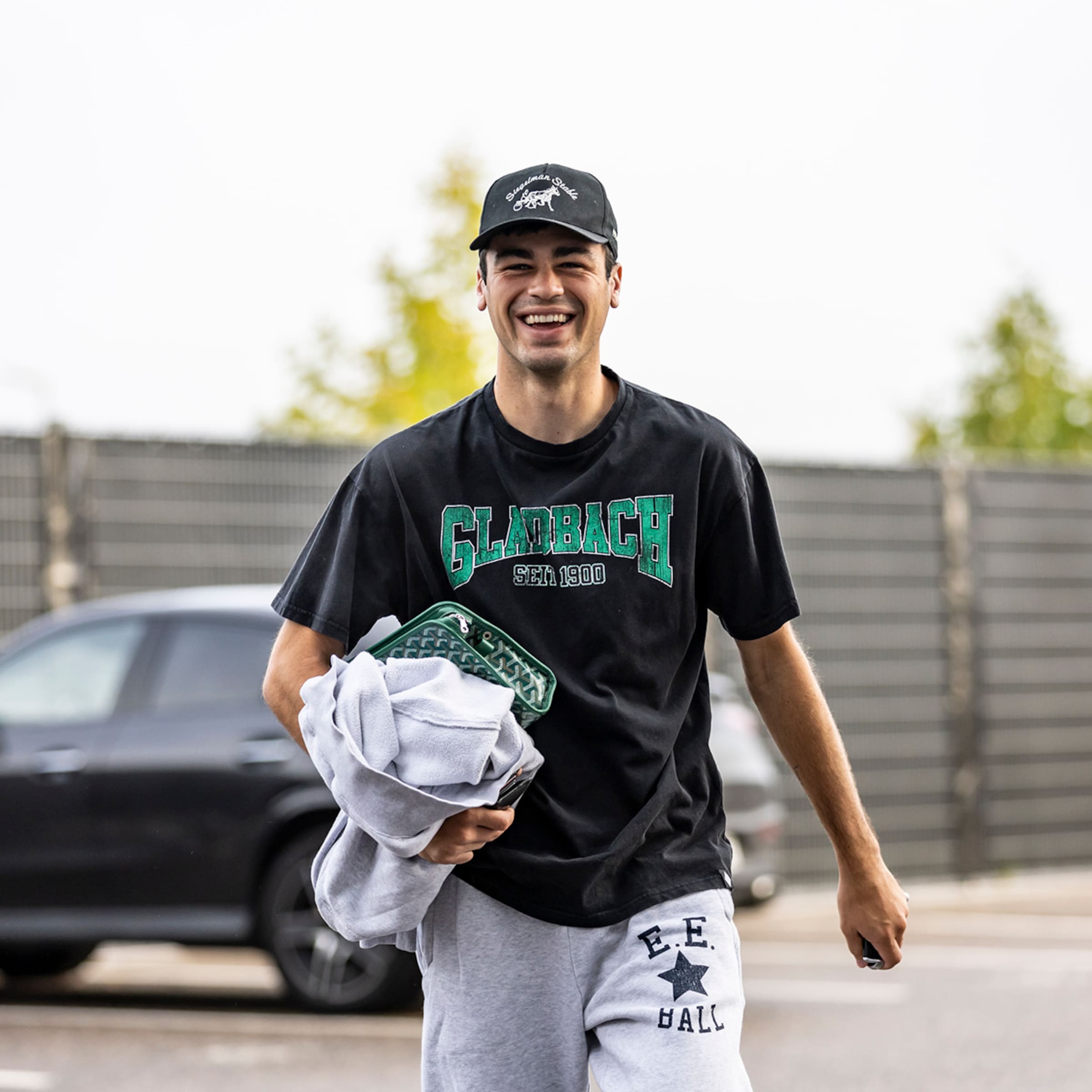 Lächelnder Mann mit Kappe, Gladbach-Shirt und Jogginghose geht mit Pullover und gemusterter grüner Tasche an einem Auto vorbei.