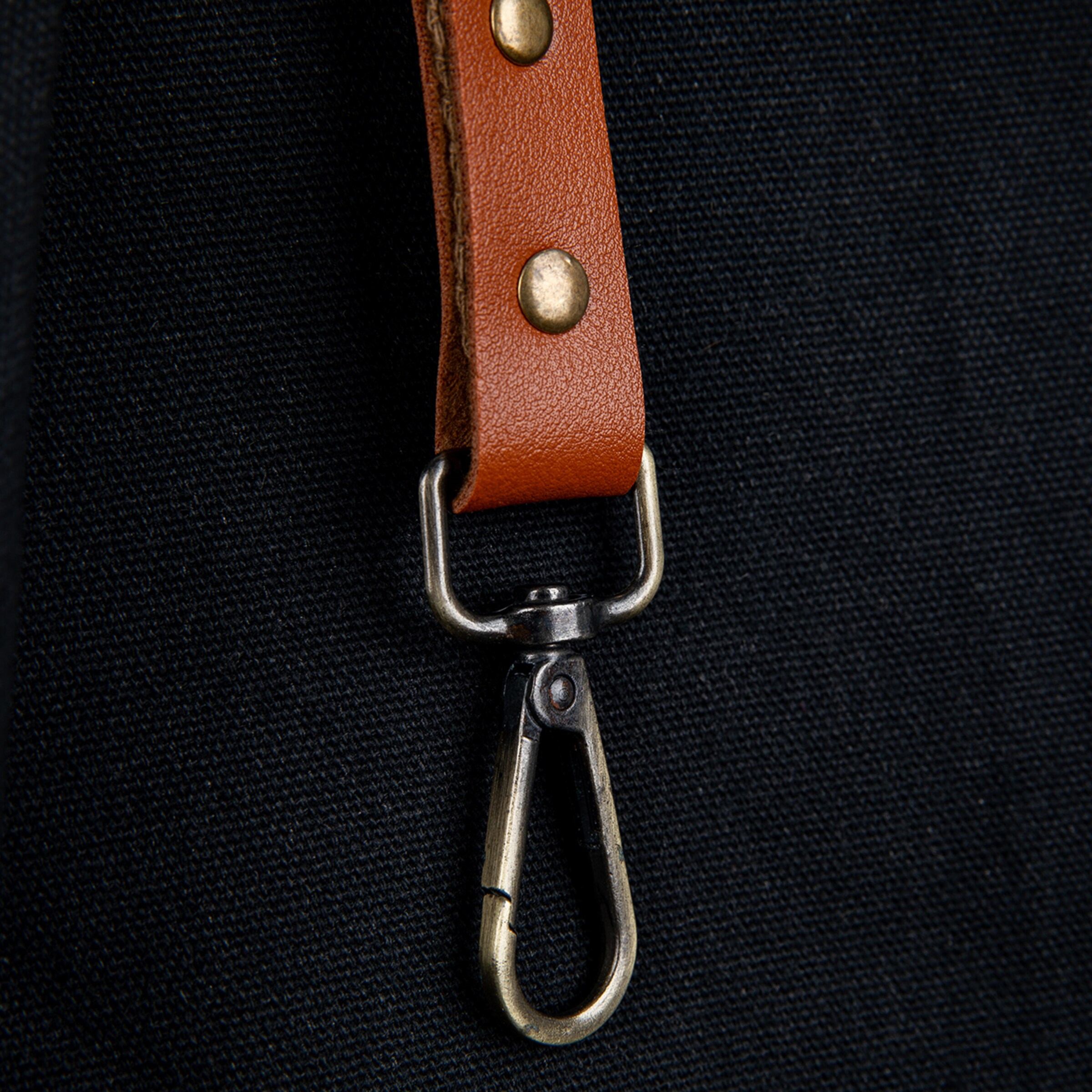 Close-up of a brown leather strap with two bronze-colored rivets, attached to a dark, woven fabric, featuring a bronze-colored swivel clasp at its end.