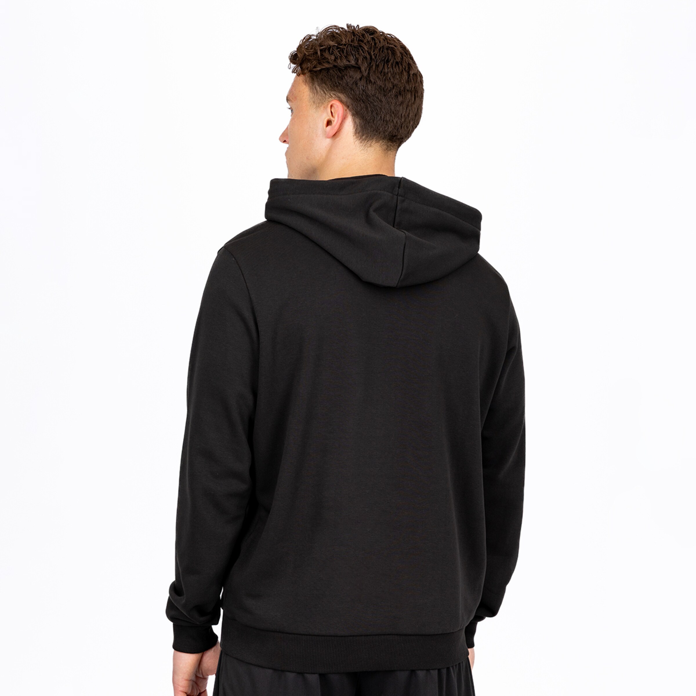 Rückansicht eines Mannes mit lockigem braunem Haar, der über seine linke Schulter blickt und einen schlichten schwarzen Kapuzenpullover sowie eine schwarze Jogginghose vor einem weißen Hintergrund trägt.