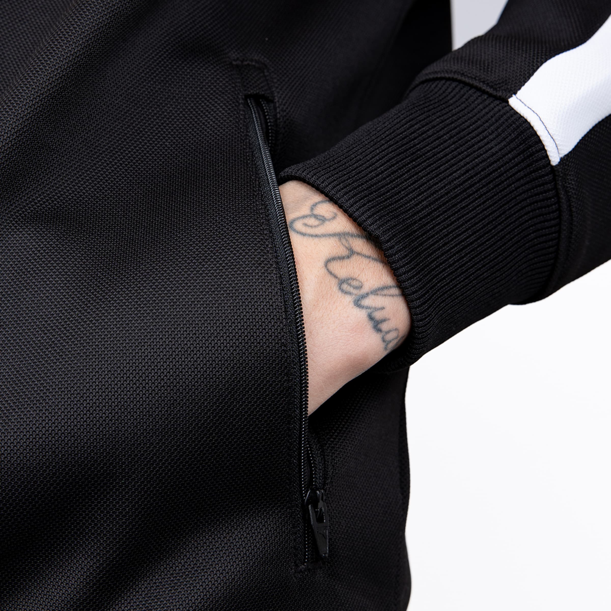 Nahaufnahme einer schwarzen Trainingsjacke mit weißem Ärmelstreifen. Eine tätowierte Hand steckt in einer seitlichen Reißverschlusstasche.