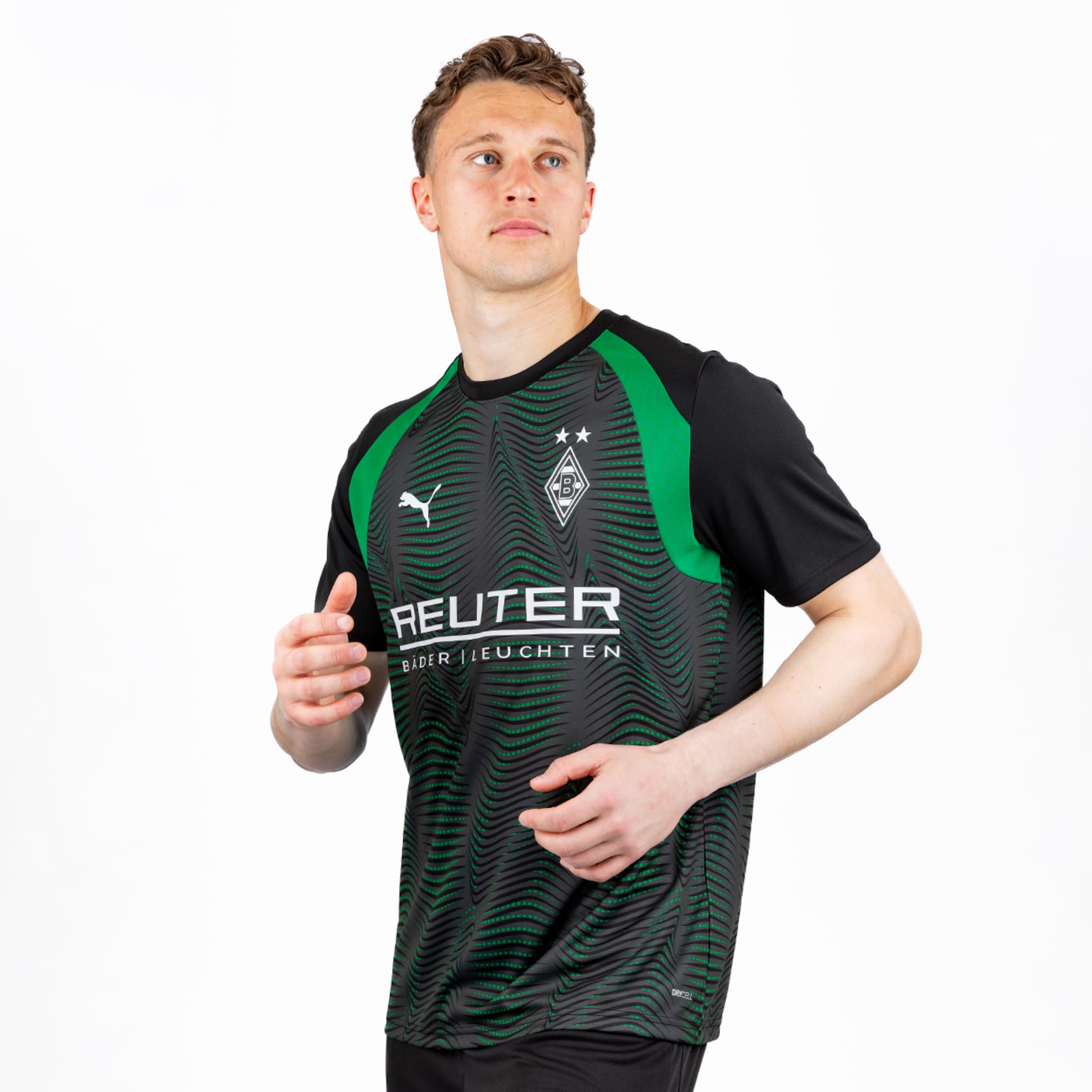 Ein junger Mann trägt ein schwarz-grünes Fußballtrikot mit einem wellenförmigen Muster, dem Puma-Logo, dem Vereinswappen und dem Sponsor "REUTER". Er blickt nach oben rechts, die Hände leicht erhoben.