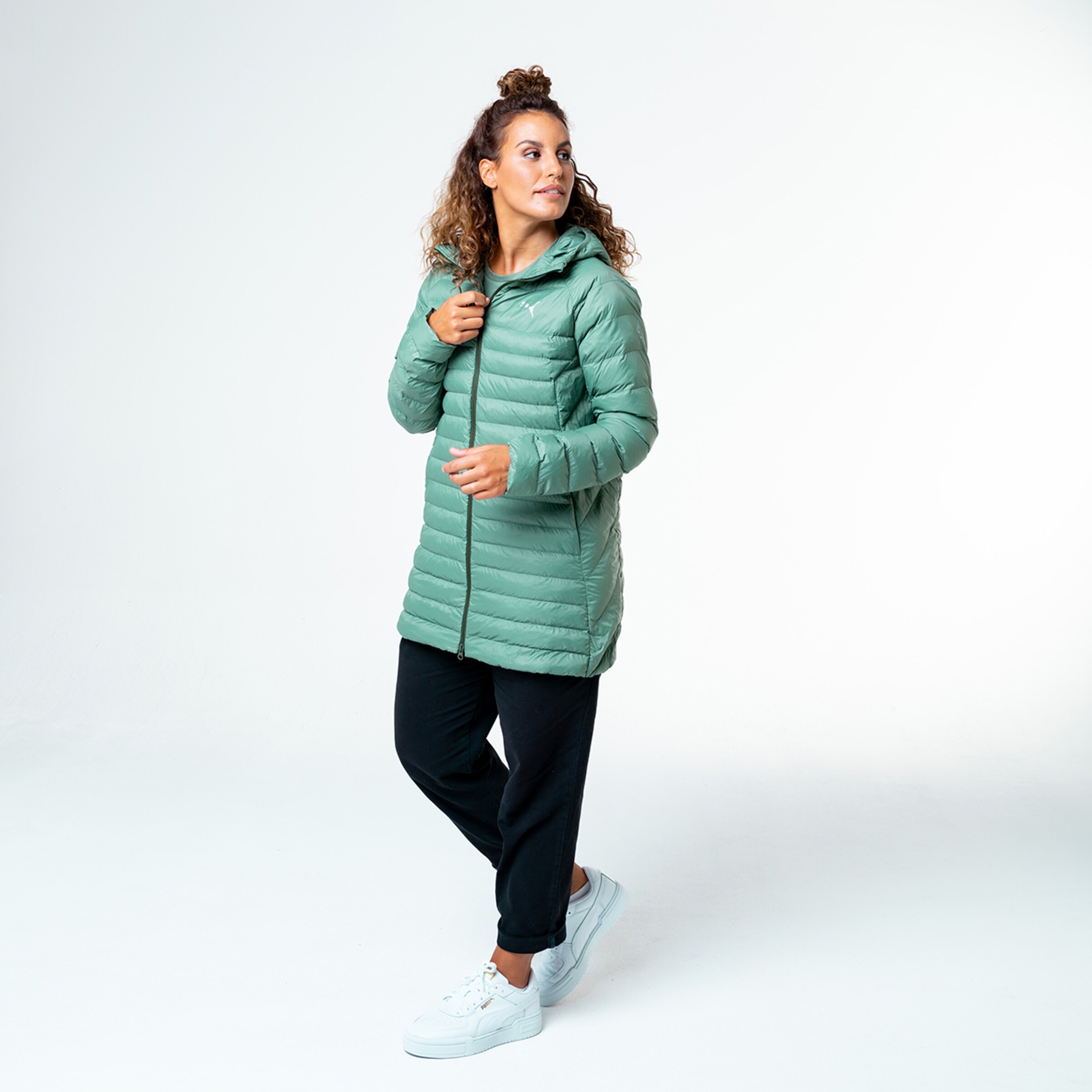 Frau in langem mintgrünem Steppjacke und schwarzen Hosen steht in Pose.