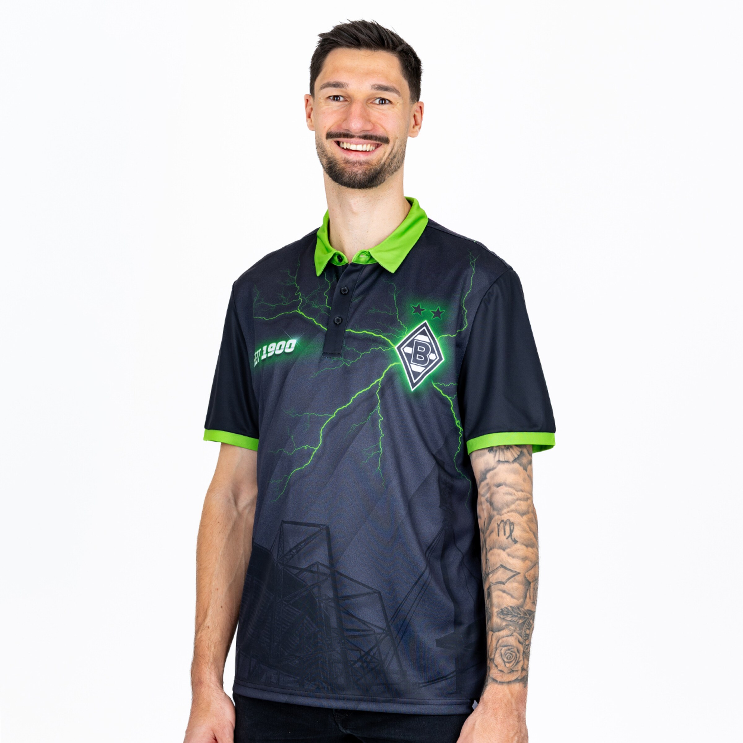 Lächelnder Mann im schwarzen Borussia Mönchengladbach Polo-Shirt mit grünen Blitzen, Logo und Ärmeltattoo vor weißem Hintergrund.