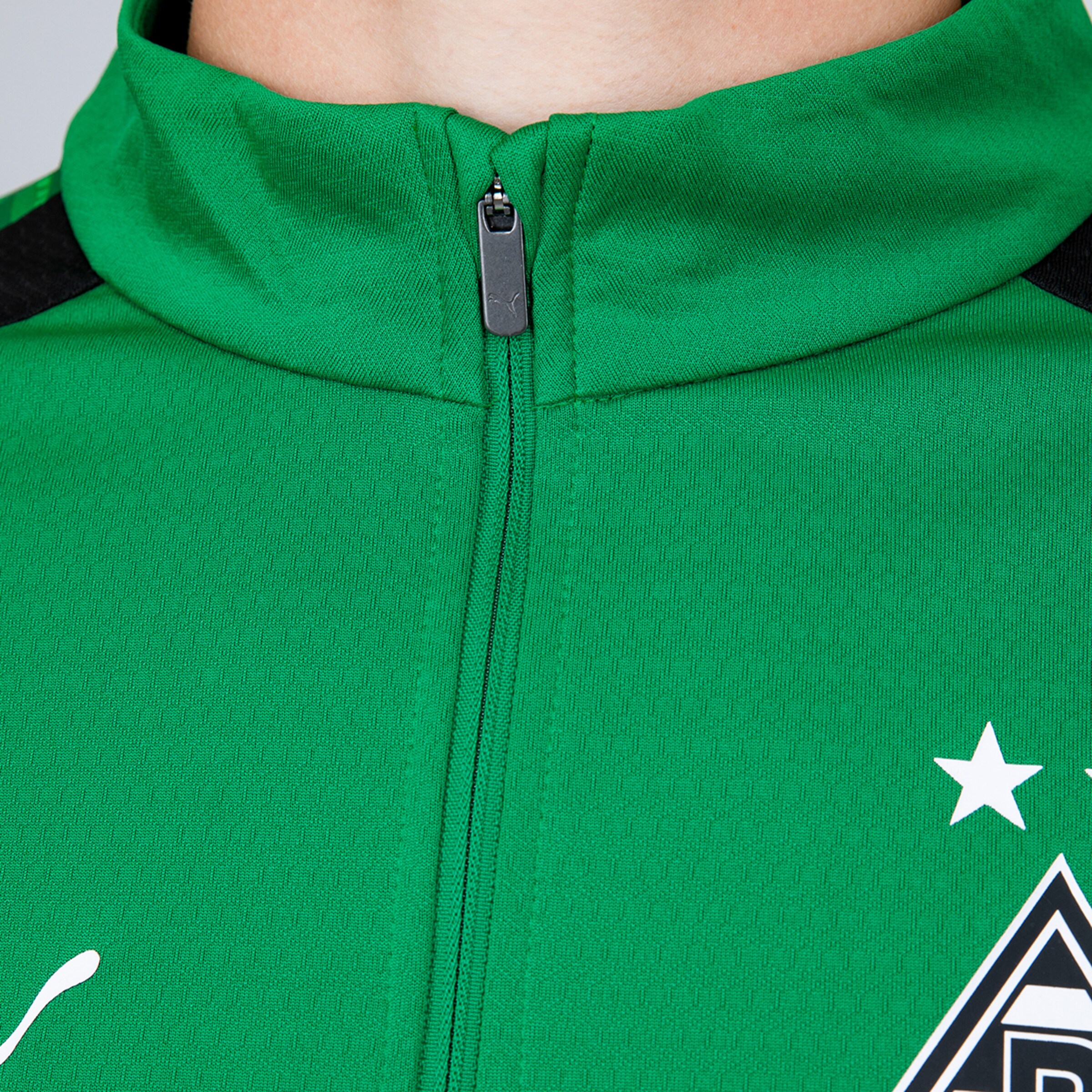 Nahaufnahme einer grünen Borussia Mönchengladbach Fußballjacke mit Stehkragen. Sie zeigt einen Reißverschluss mit PUMA-Logo, einen weißen Stern und das Vereinswappen rechts, sowie ein weiteres weißes PUMA-Logo links.