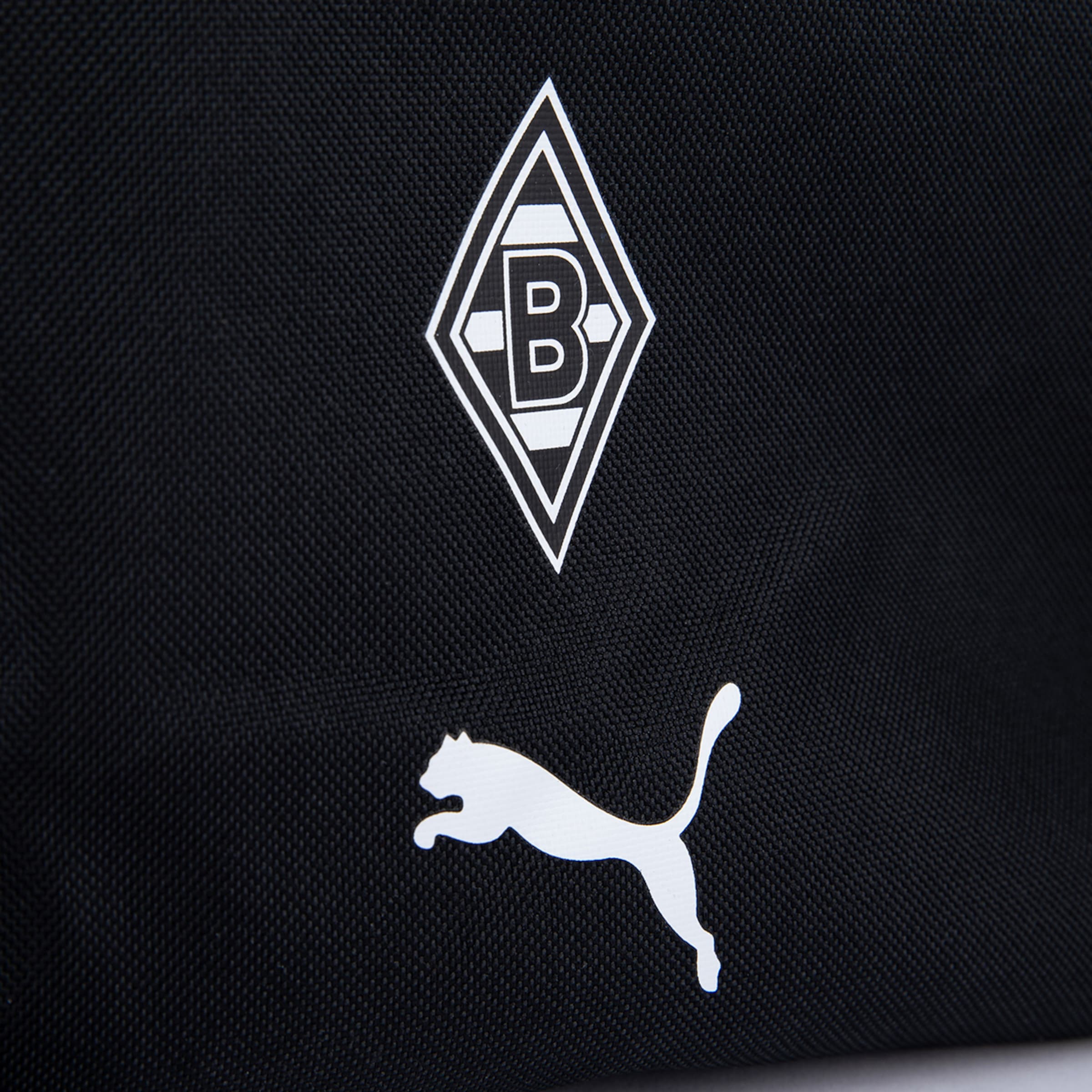 Nahaufnahme einer schwarzen Textiloberfläche mit zwei weißen Logos: Oben das Rautenlogo von Borussia Mönchengladbach mit einem 'B' und zwei Hexagonen, darunter das springende Katzenlogo von PUMA.