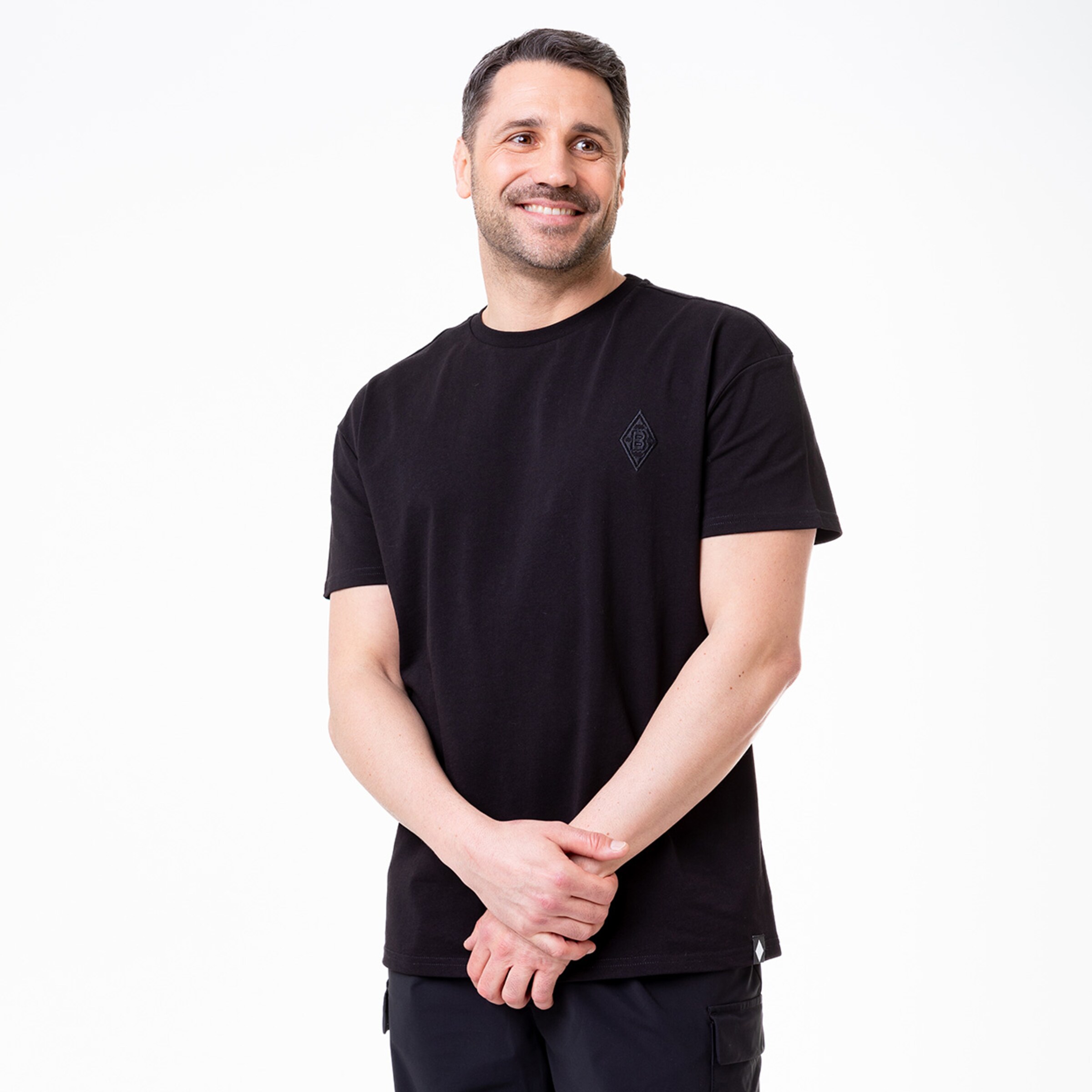 Mann mit lächelndem Gesicht in schwarzem T-Shirt steht mit verschränkten Armen.