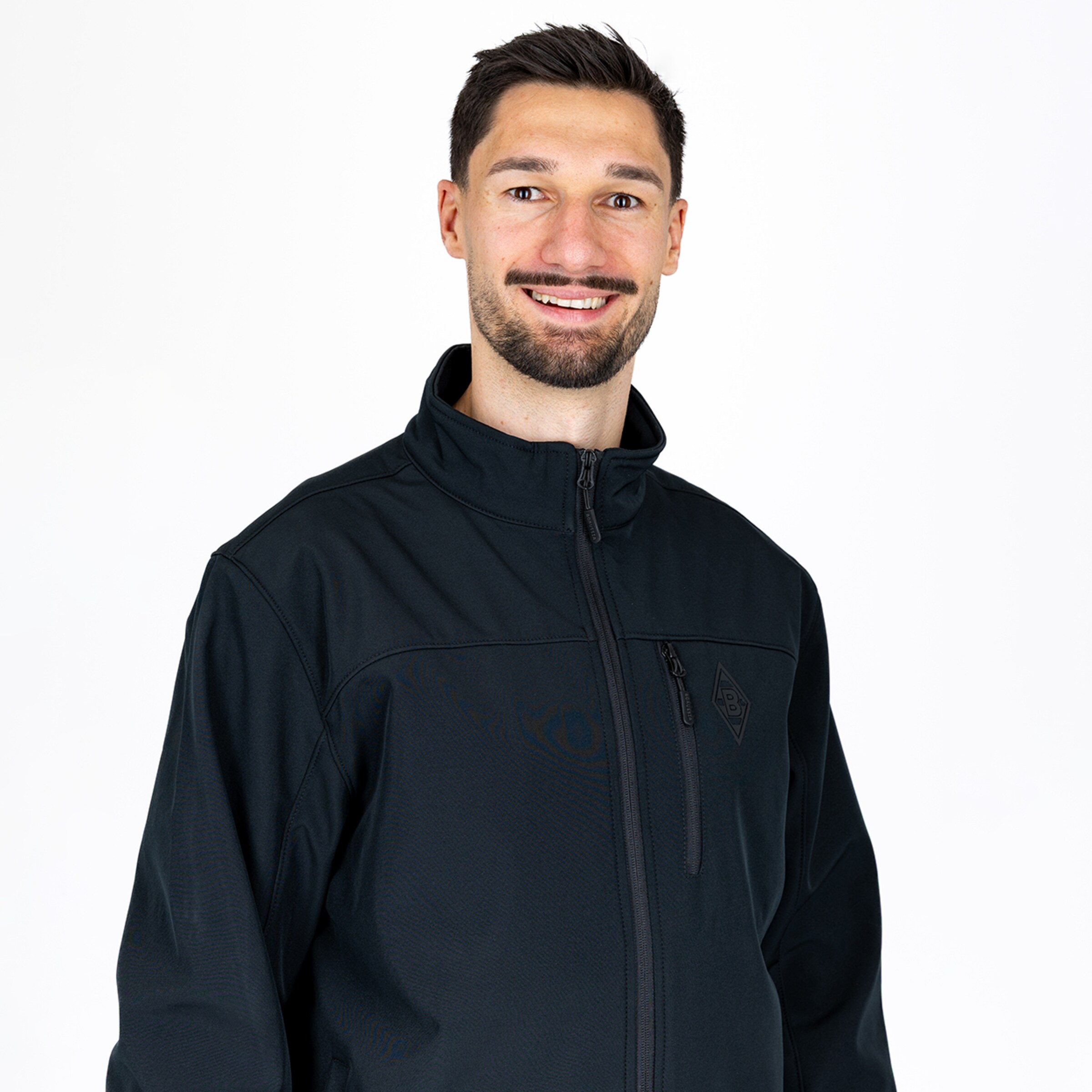 Porträt eines lächelnden Mannes mit Bart in einer schwarzen Borussia-Mönchengladbach-Jacke vor weißem Hintergrund.