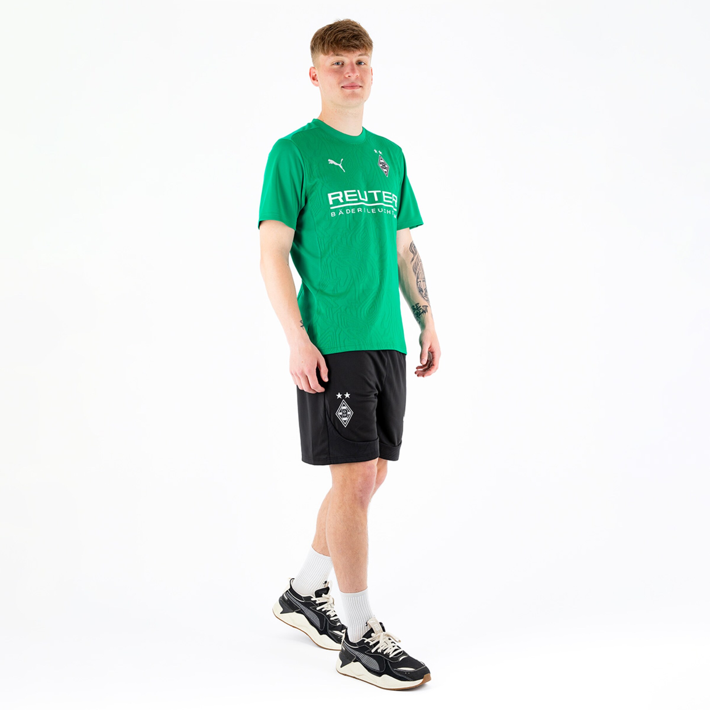 Junger Mann in grünem Sportshirt und schwarzen Shorts steht aufrecht.